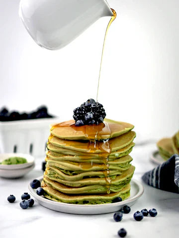 A towering stack of fluffy matcha pancakes topped with fresh blackberries and blueberries, drizzled with golden syrup for a vibrant and delicious breakfast