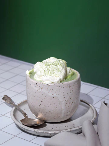 A cozy ceramic mug filled with creamy matcha latte topped with a generous dollop of whipped coconut cream and a sprinkle of matcha powder, set against a modern green backdrop.