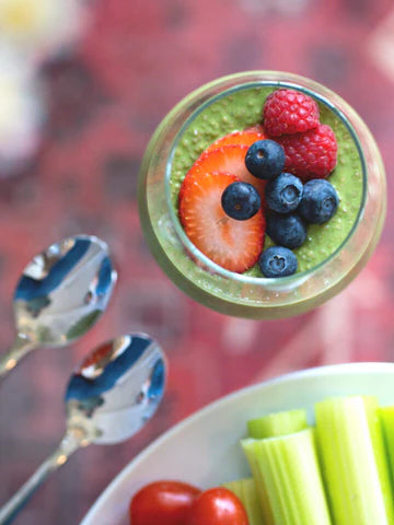 A jar of bright green matcha chia pudding layered with colorful fresh berries, accompanied by crisp celery sticks and cherry tomatoes for a healthy, balanced snack.