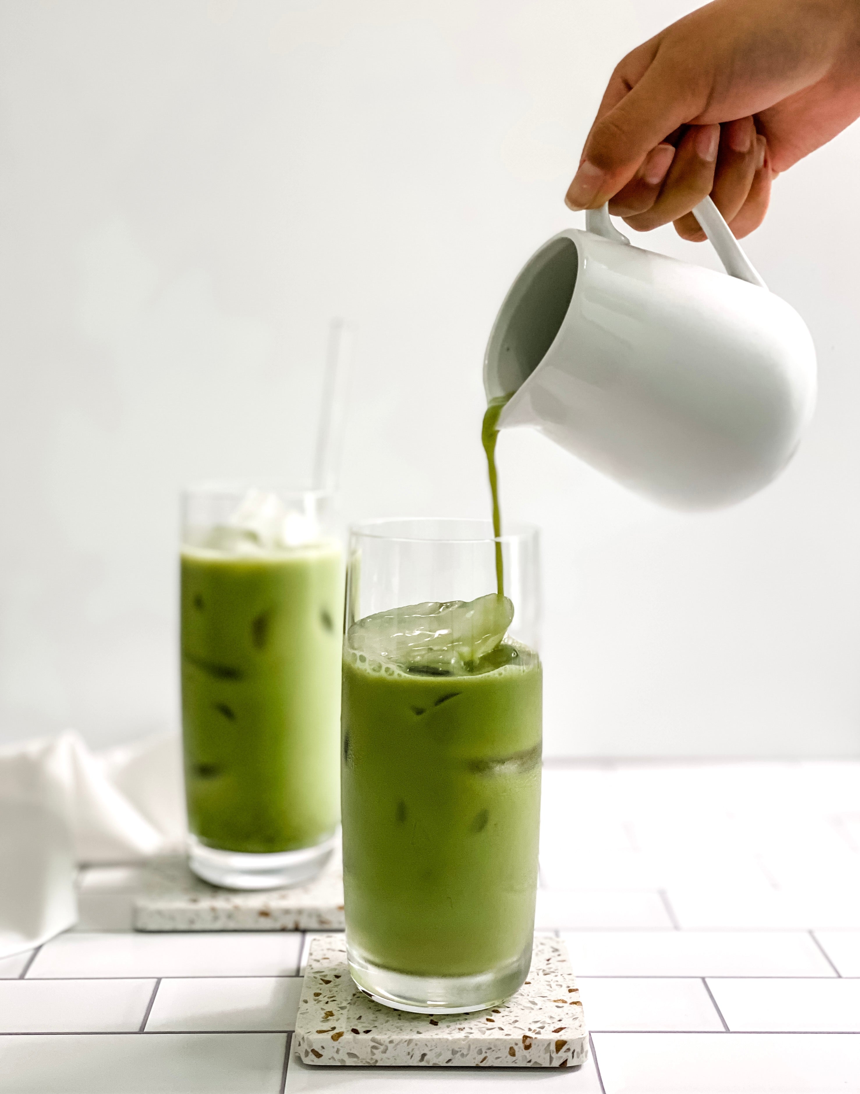 Two tall glasses of iced matcha latte on terrazzo coasters, one being topped with freshly whisked matcha poured from a white pitcher, creating a refreshing and vibrant green drink perfect for warm days.