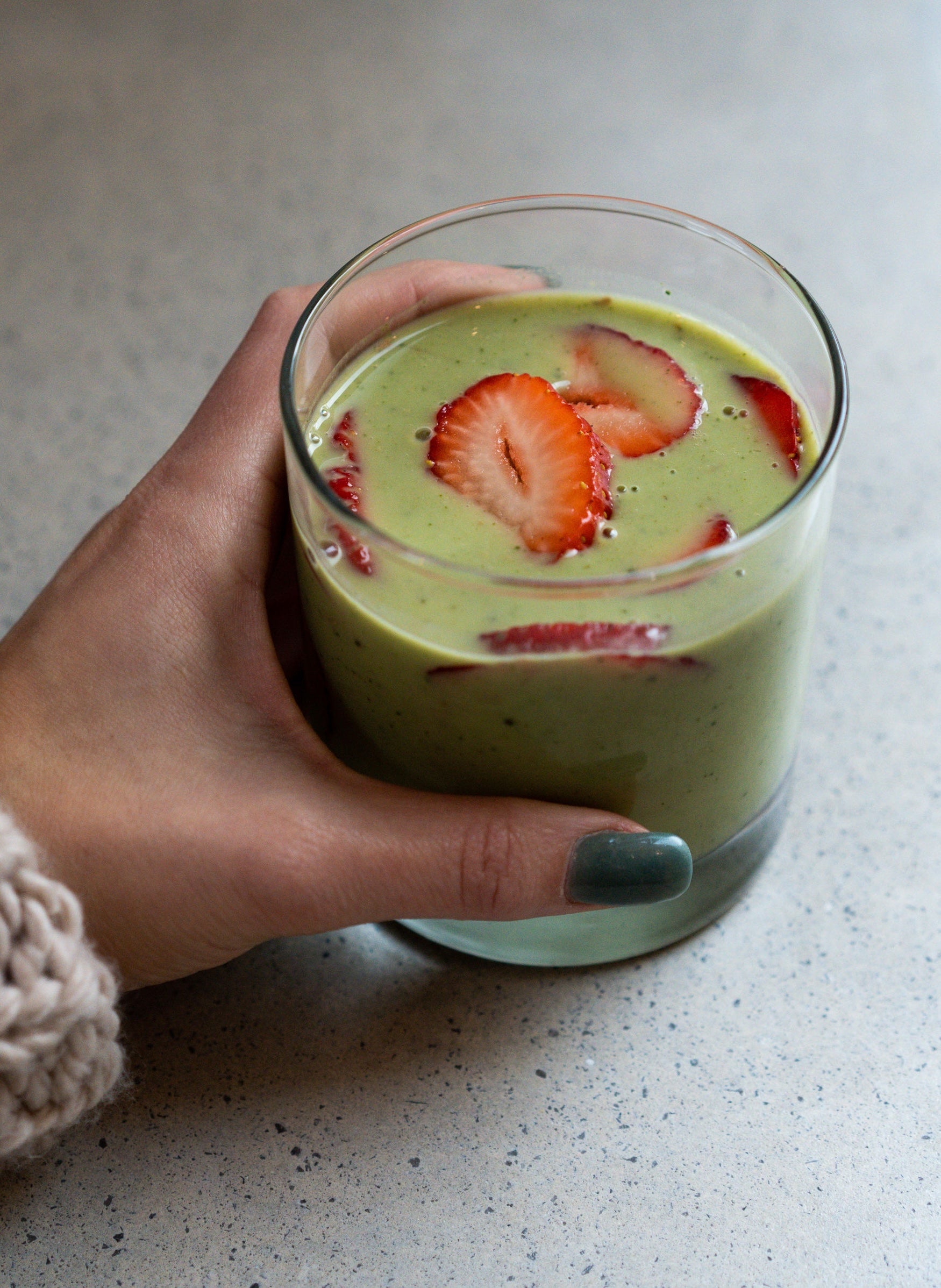 A hand holding a glass of creamy matcha smoothie topped with vibrant slices of fresh strawberries, creating a colorful and nourishing treat perfect for any time of the day.