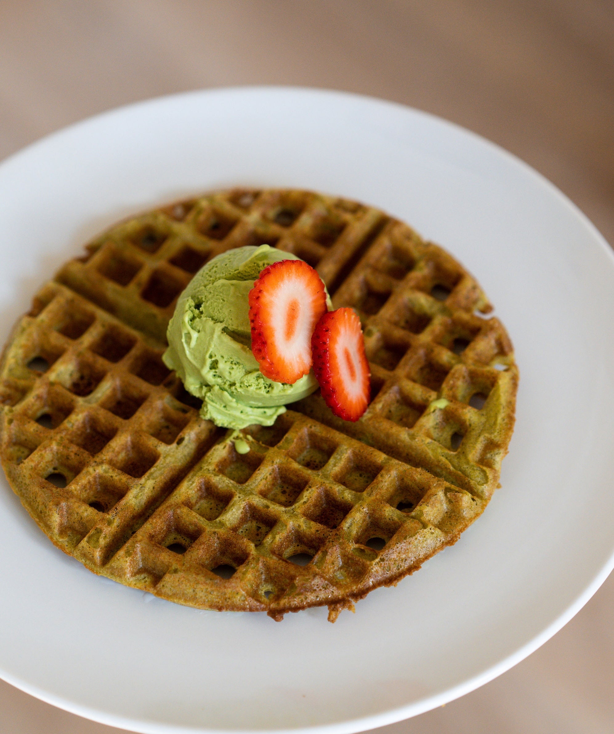 A golden-brown matcha waffle served on a white plate, topped with a scoop of creamy matcha ice cream and fresh strawberry slices for a colorful and indulgent breakfast or dessert.