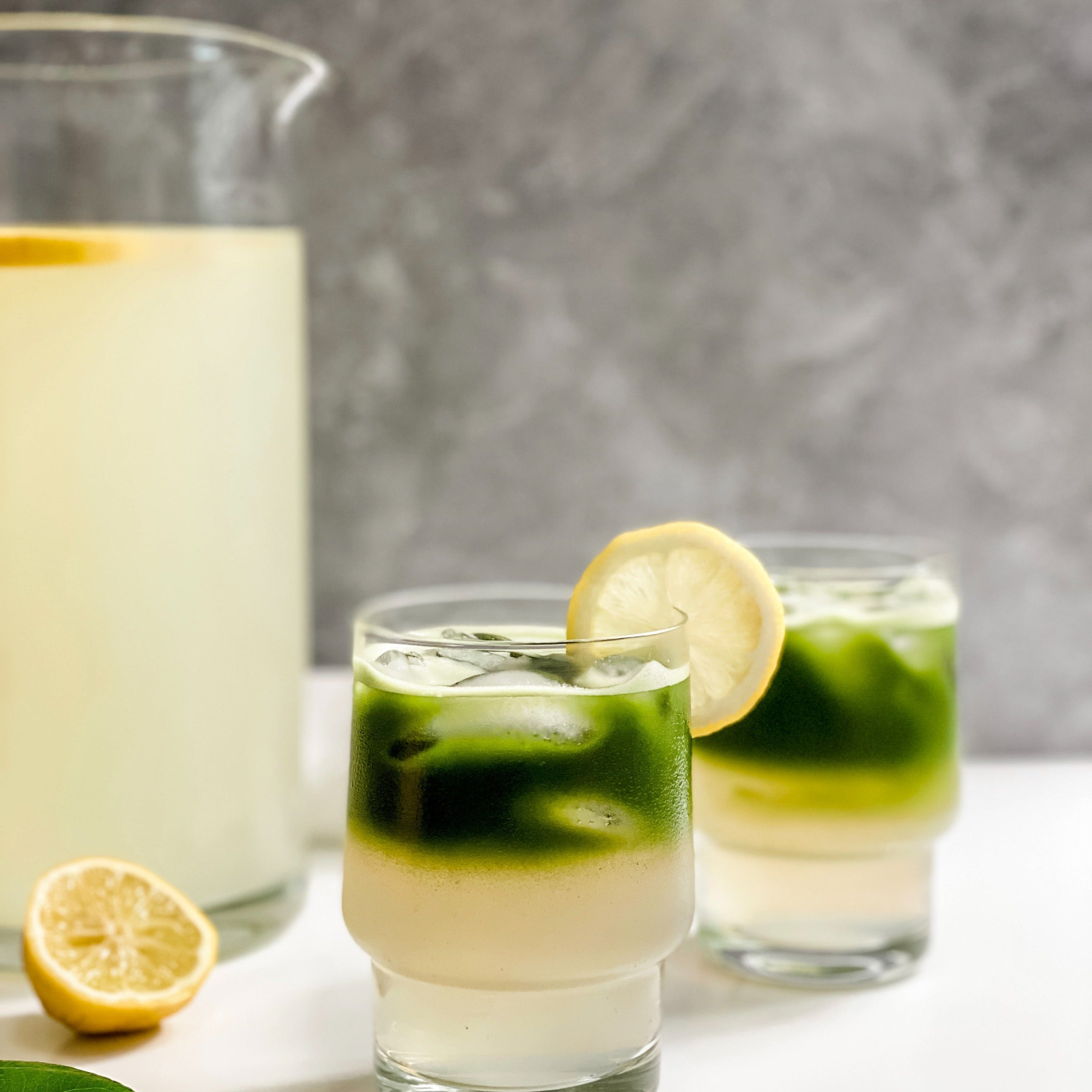 Two glasses of vibrant matcha lemonade served over ice, with layers of green matcha and zesty lemonade topped with lemon slices, set against a backdrop featuring a pitcher of fresh lemonade and a sliced lemon for garnish