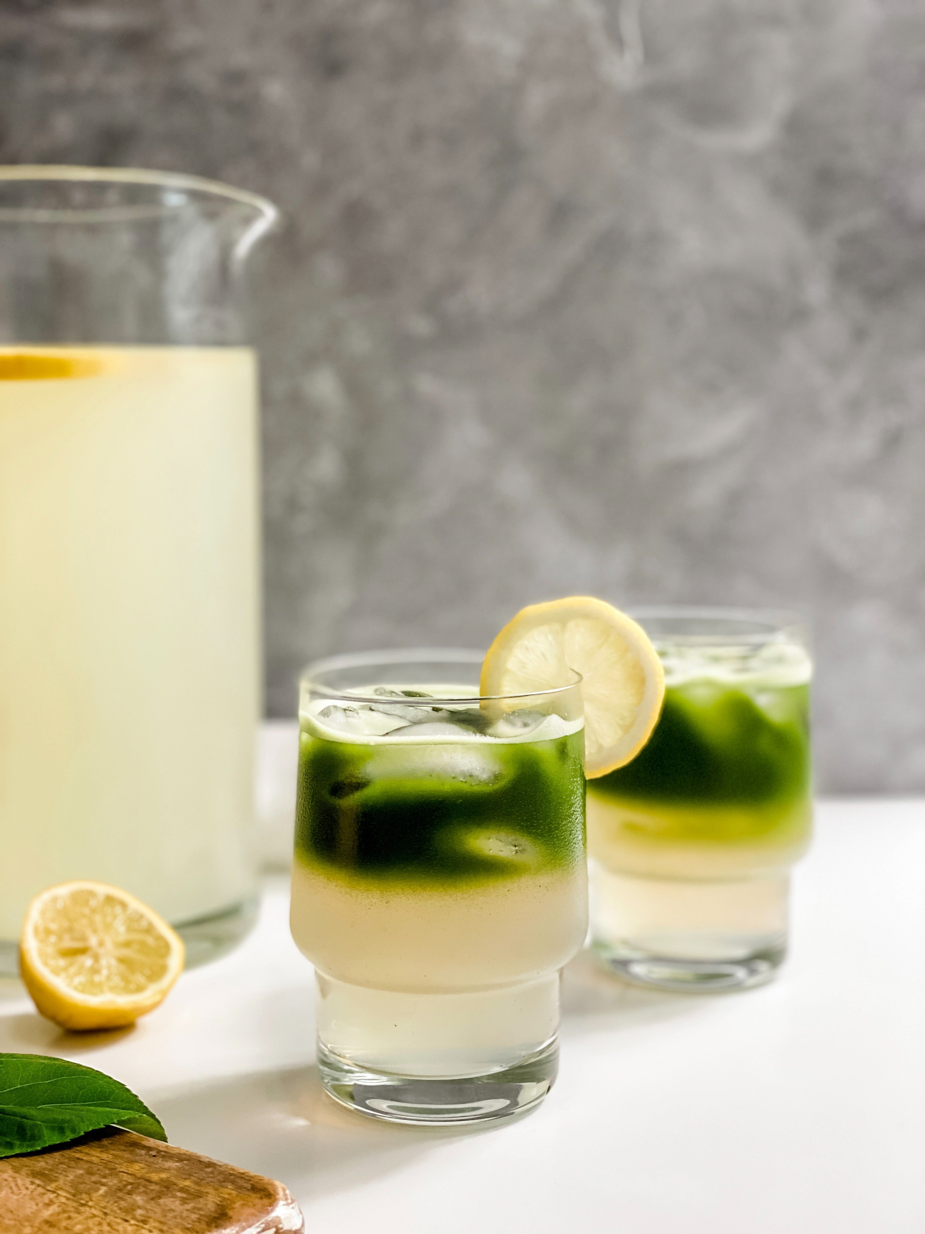 Two glasses of vibrant matcha lemonade served over ice, with layers of green matcha and zesty lemonade topped with lemon slices, set against a backdrop featuring a pitcher of fresh lemonade and a sliced lemon for garnish