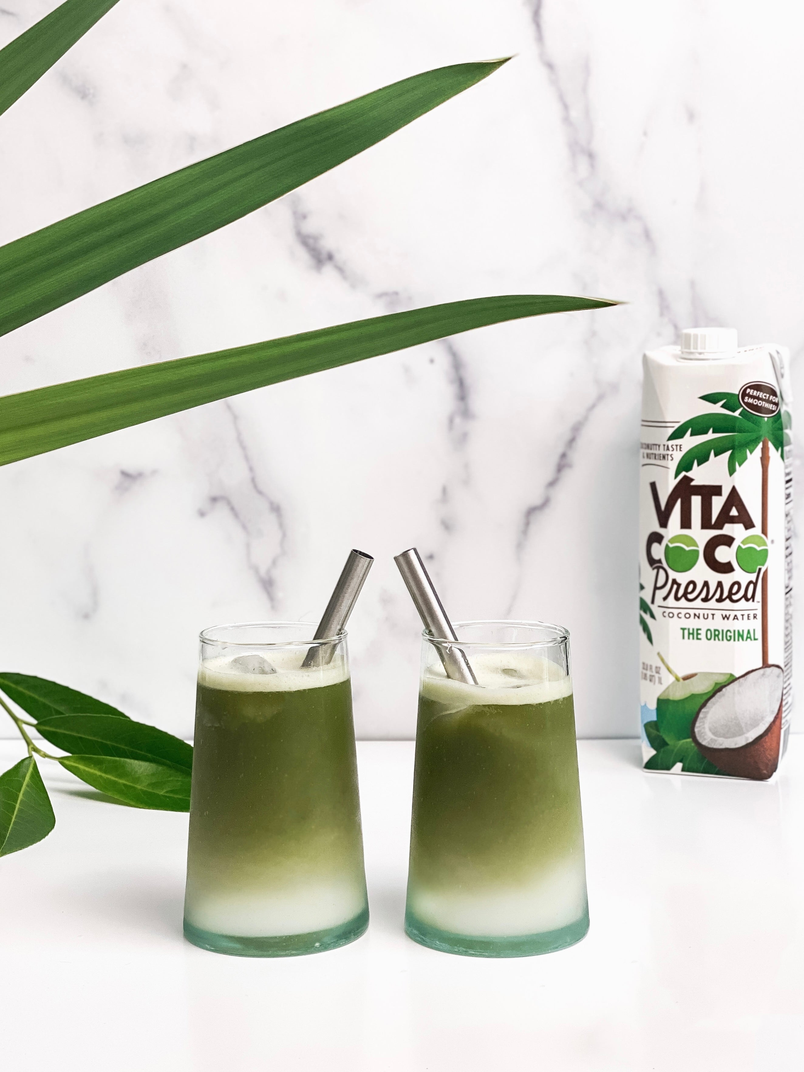 Two glasses of vibrant coconut water matcha, layered with creamy coconut water and green matcha, served over ice with stainless steel straws, set against a marble backdrop with tropical leaves and a Vita Coco Pressed carton in the background.