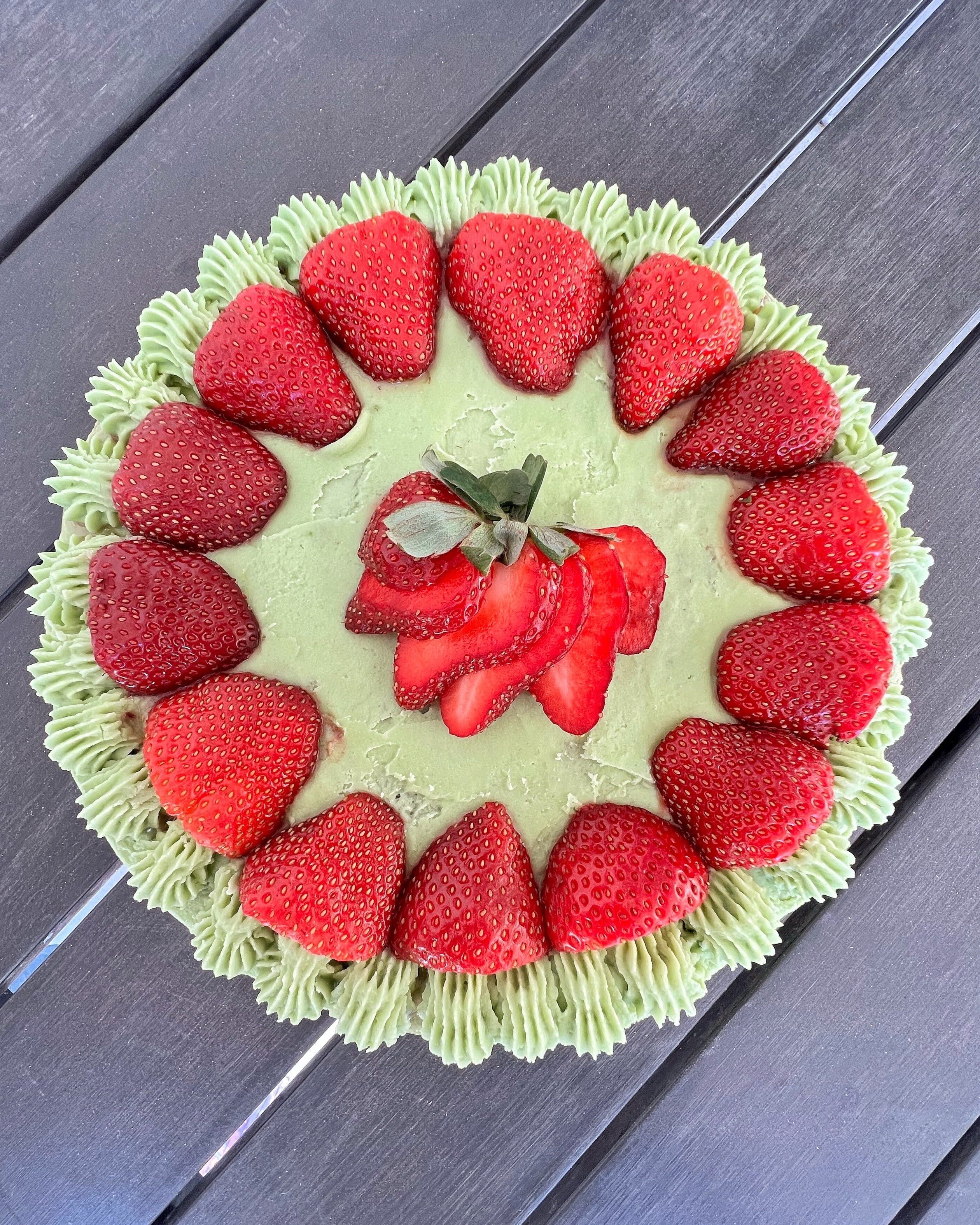 A beautifully decorated matcha cake topped with vibrant green matcha frosting, surrounded by fresh whole strawberries along the edge and layered strawberry slices in the center, creating a stunning dessert centerpiece.