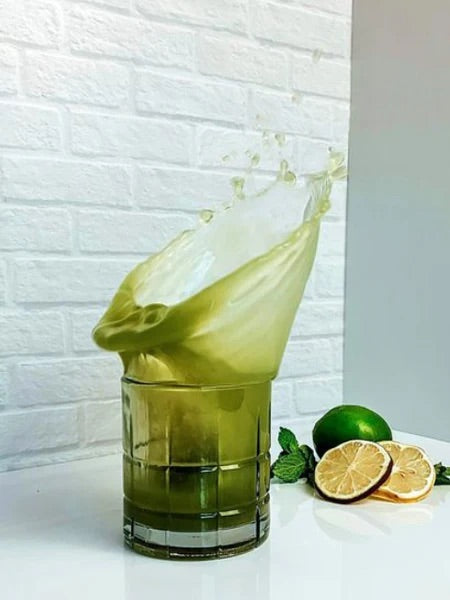 A dynamic shot of a Matcha Nude Highball in a clear glass, capturing the splash of the matcha cocktail as it spills over the rim. Fresh lime slices, a whole lime, and mint leaves are placed on a white surface beside the glass.