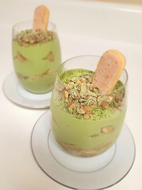 Two individual servings of matcha tiramisu layered in glass cups, topped with crushed pistachios, a sprinkle of matcha powder, and a ladyfinger biscuit. 