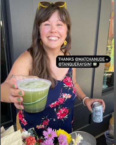 The owner of Matcha Nude holds out a class of a Matcha Gin Collins towards the camera. She is wearing a navy blue and floral tank top with flowers in the foreground.