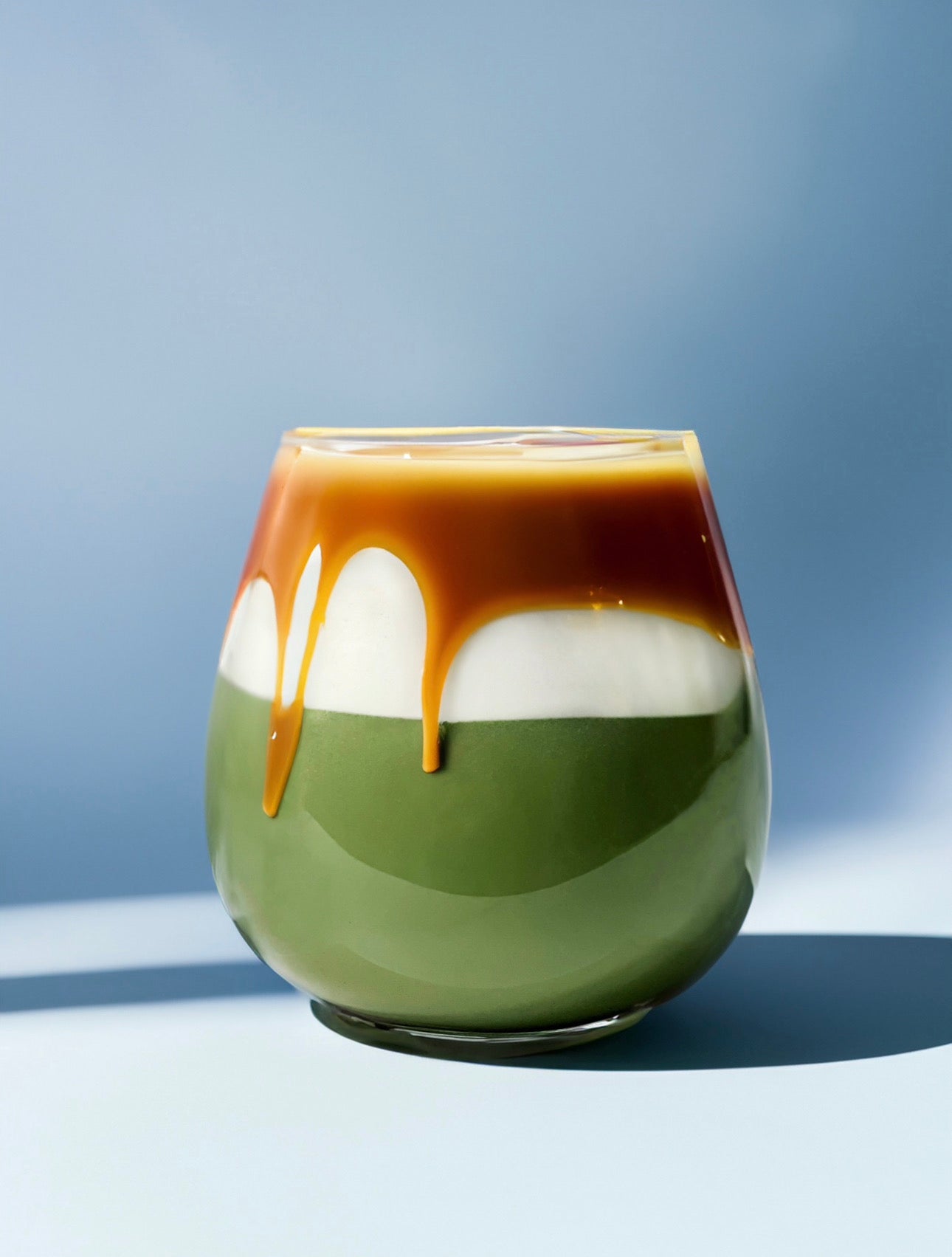  A stunning salted caramel matcha latte in a clear glass, showcasing vibrant green matcha at the base, a creamy white milk layer, and rich caramel sauce dripping elegantly over the top against a soft blue background. 🍵