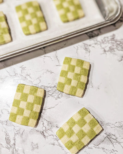 Checkerboard matcha cookies with a perfect green and white pattern, beautifully arranged on a marble countertop, with a baking tray in the background showcasing freshly baked treats. 🍵✨
