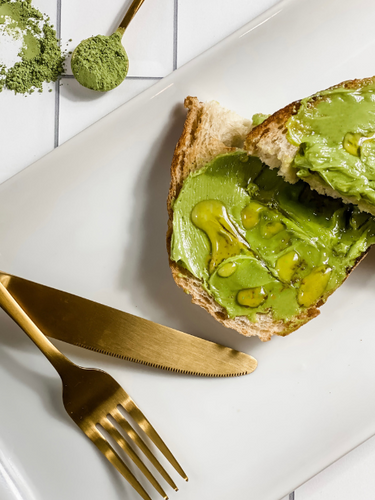 A slice of crusty bread generously spread with vibrant green matcha honey butter, drizzled with golden honey, and paired with elegant gold flatware on a sleek white plate, with a touch of matcha powder in the background for a perfect pop of color. 🍵✨
