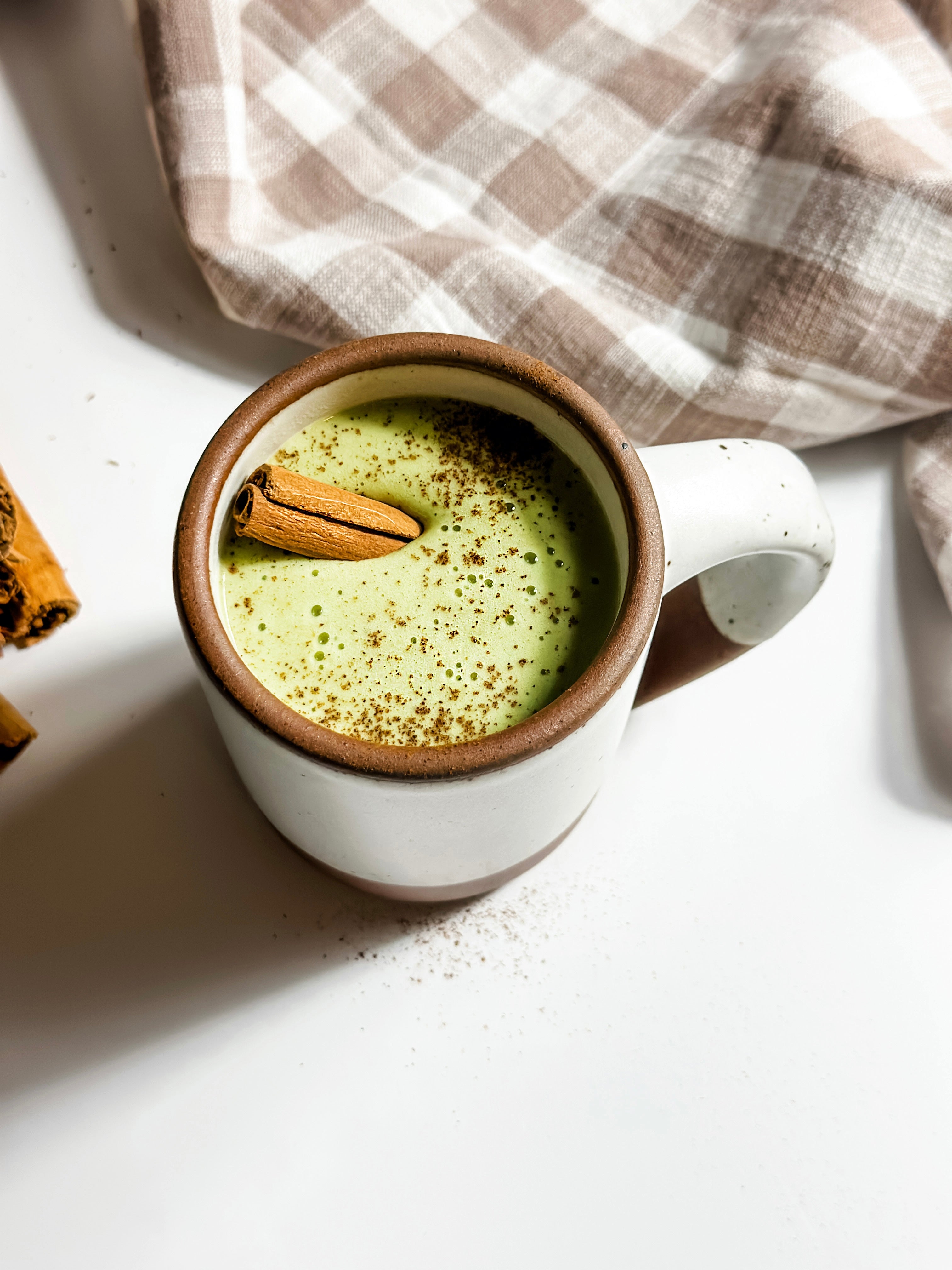  A cozy cinnamon matcha latte served in a rustic white mug with a speckled brown rim, topped with a cinnamon stick and a sprinkle of cinnamon powder, set against a soft plaid napkin for ultimate warm and inviting vibes. 🍵✨