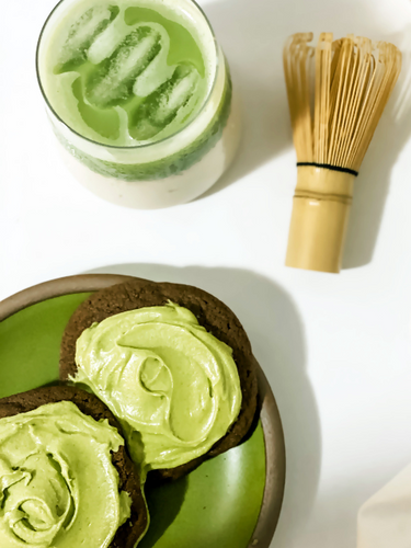 Two chocolate cookies topped with a silky, vibrant green matcha French maple buttercream, paired with a creamy iced matcha latte and a traditional bamboo whisk, creating a decadent and elegant treat. 🍵✨