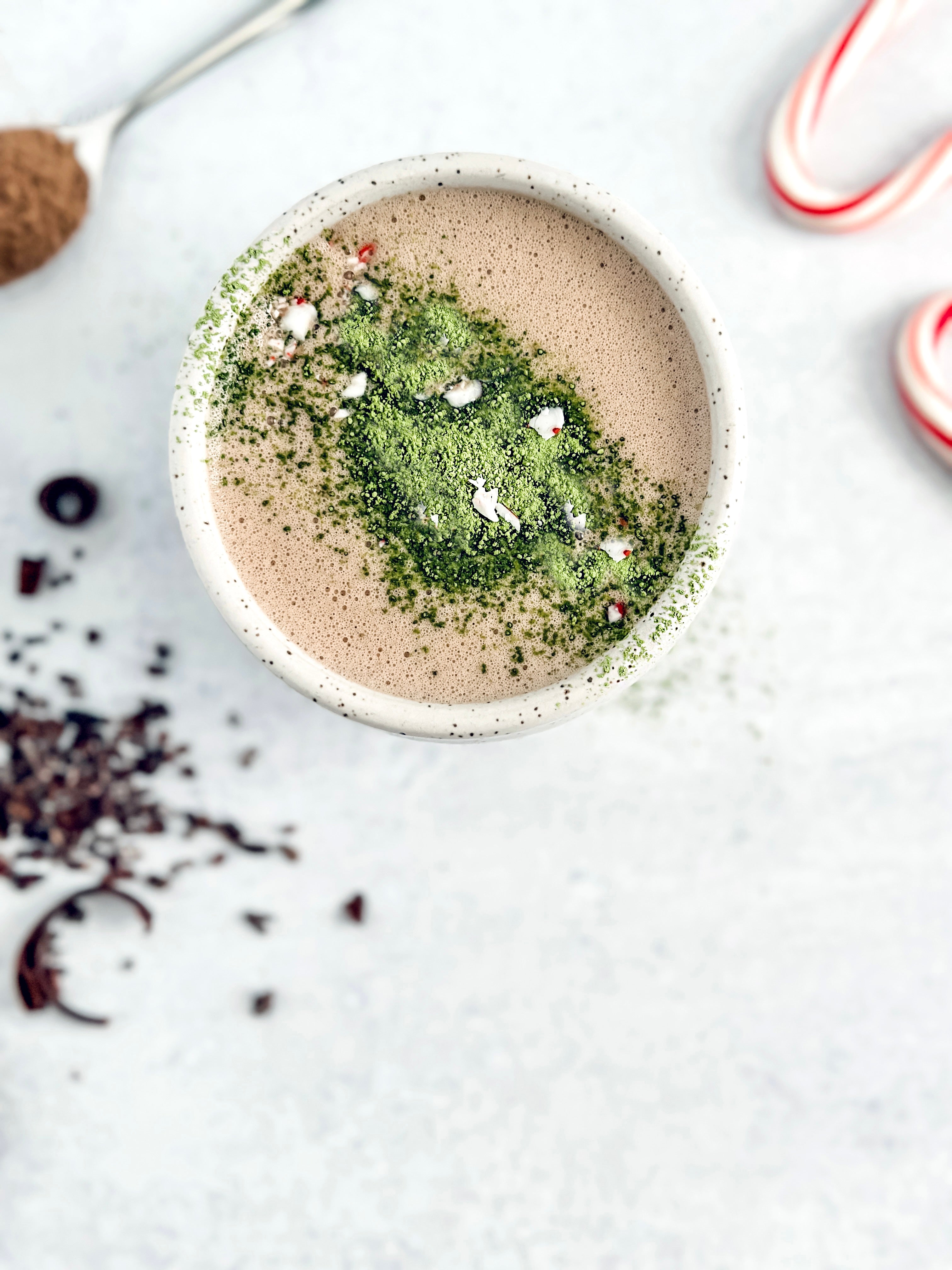 A creamy peppermint cacao matcha latte served in a speckled ceramic mug, topped with vibrant green matcha powder and crushed peppermint pieces, surrounded by festive candy canes and cacao nibs for a cozy holiday treat. 