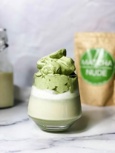 A vibrant Matcha Dalgona latte with fluffy, whipped matcha topping sitting atop a creamy milk base, served in a clear glass against a marble surface, with Matcha Nude packaging and a milk bottle in the background for a cozy yet elegant setup.