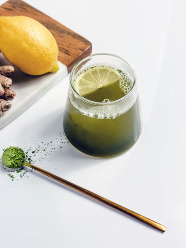 A vibrant Matcha Zinger hot drink in a clear glass with a lemon slice on top, paired with fresh lemon, ginger, and a golden spoon holding matcha powder. 🍵🍋✨