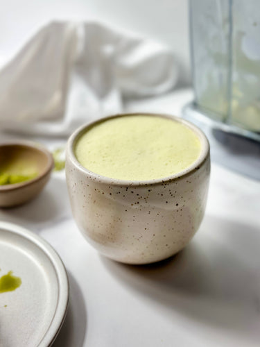 A creamy bone broth matcha latte served in a speckled ceramic mug, with a warm, frothy top, set against a minimalist backdrop featuring subtle earthy tones and a blender in the background. 🍵✨