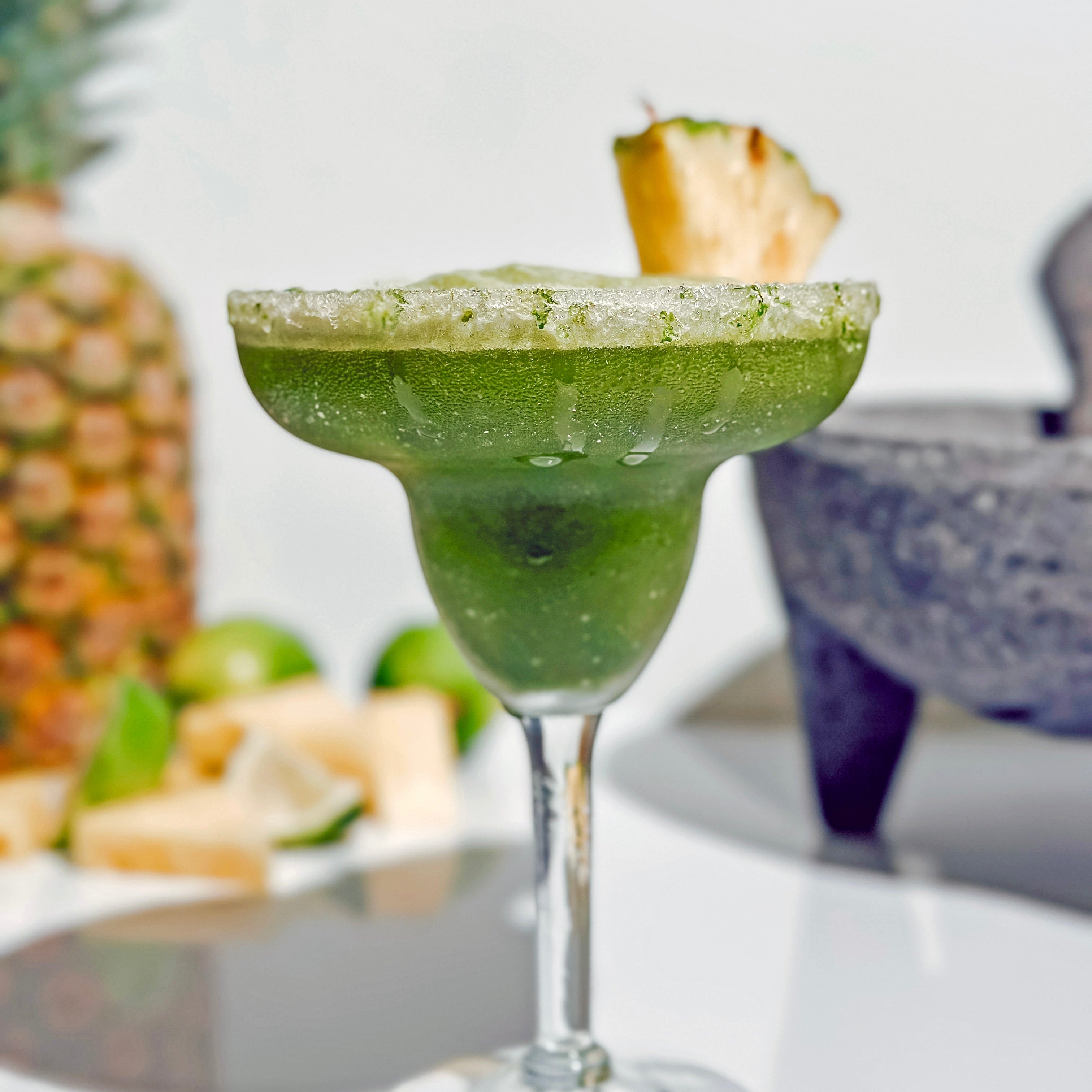  A tropical Pineapple Matcha Margarita in a frosted glass, garnished with a fresh pineapple wedge. The drink sparkles in the sunlight, surrounded by limes and pineapple chunks for a vibrant summer vibe