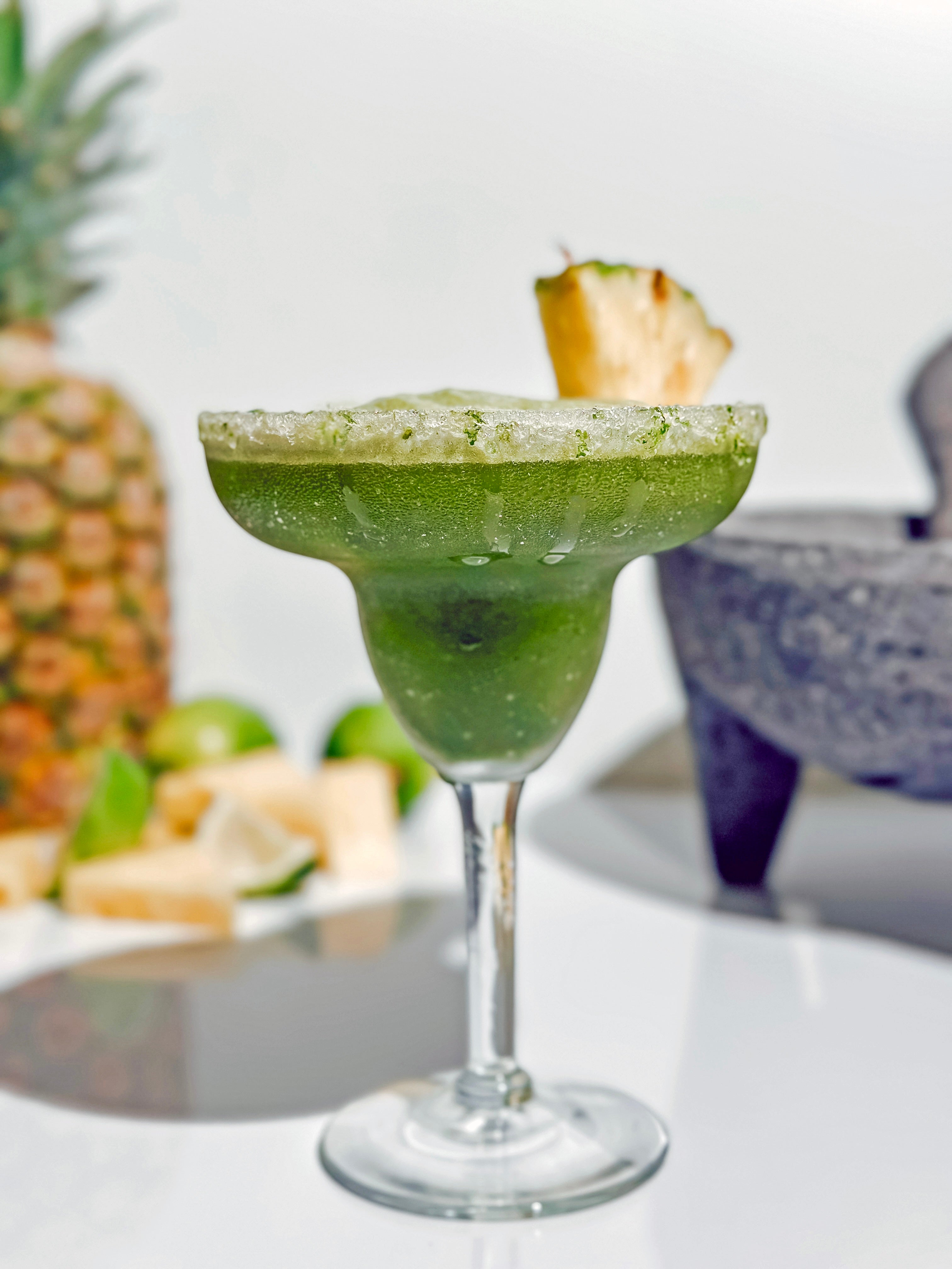  A tropical Pineapple Matcha Margarita in a frosted glass, garnished with a fresh pineapple wedge. The drink sparkles in the sunlight, surrounded by limes and pineapple chunks for a vibrant summer vibe