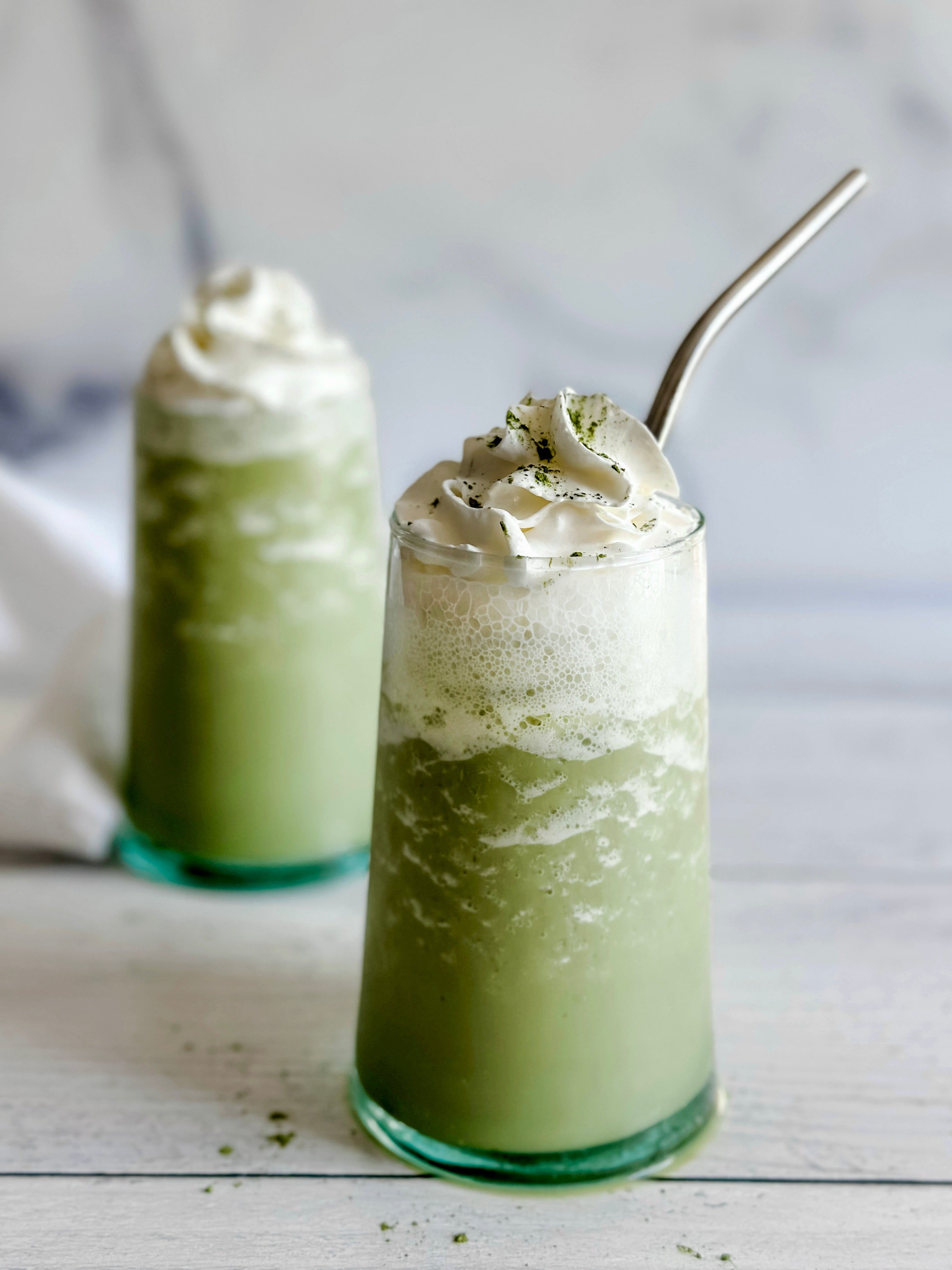 Two creamy Matcha Frappuccinos in tall glasses, topped with fluffy whipped cream and a dusting of matcha powder, with a metal straw adding a polished touch. 🍵✨