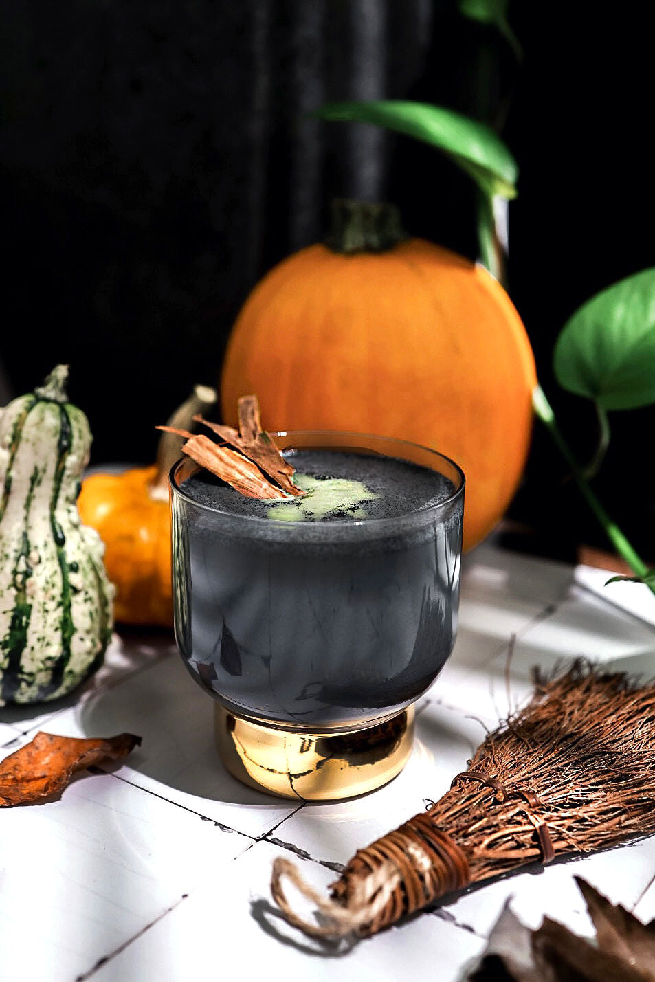 A spooky Wicked Witch Matcha Latte in a glass with a dark charcoal hue, topped with a swirl of matcha and cinnamon sticks, surrounded by pumpkins, gourds, and autumn leaves on a moody background.