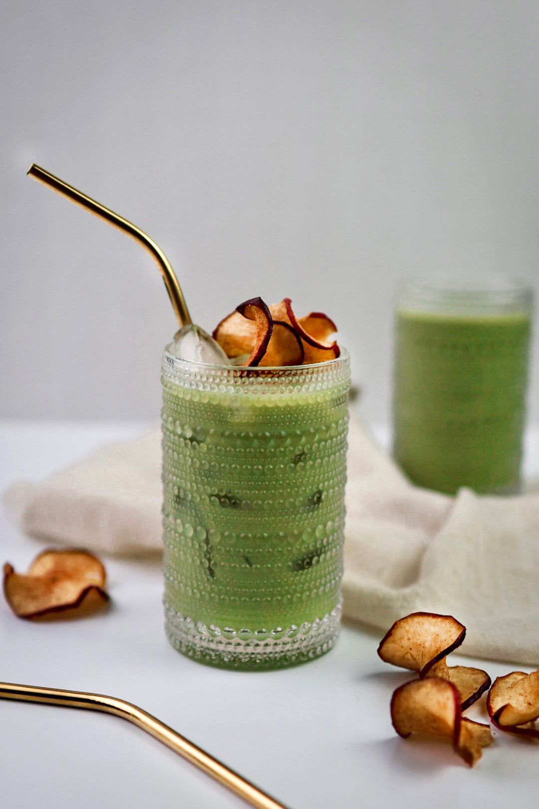A cozy Apple Cider Matcha Latte served in a textured glass, topped with dried apple slices, and paired with a gold straw, surrounded by fall-inspired decor.