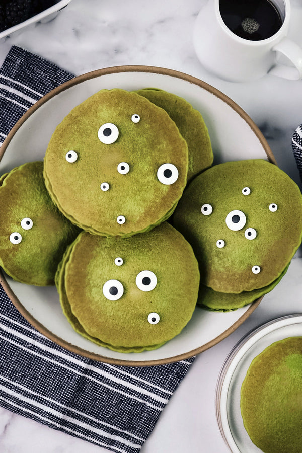 A stack of green Matcha Monster Pancakes topped with candy eyes, served on a white plate with maple syrup and a cup of coffee in the background.
