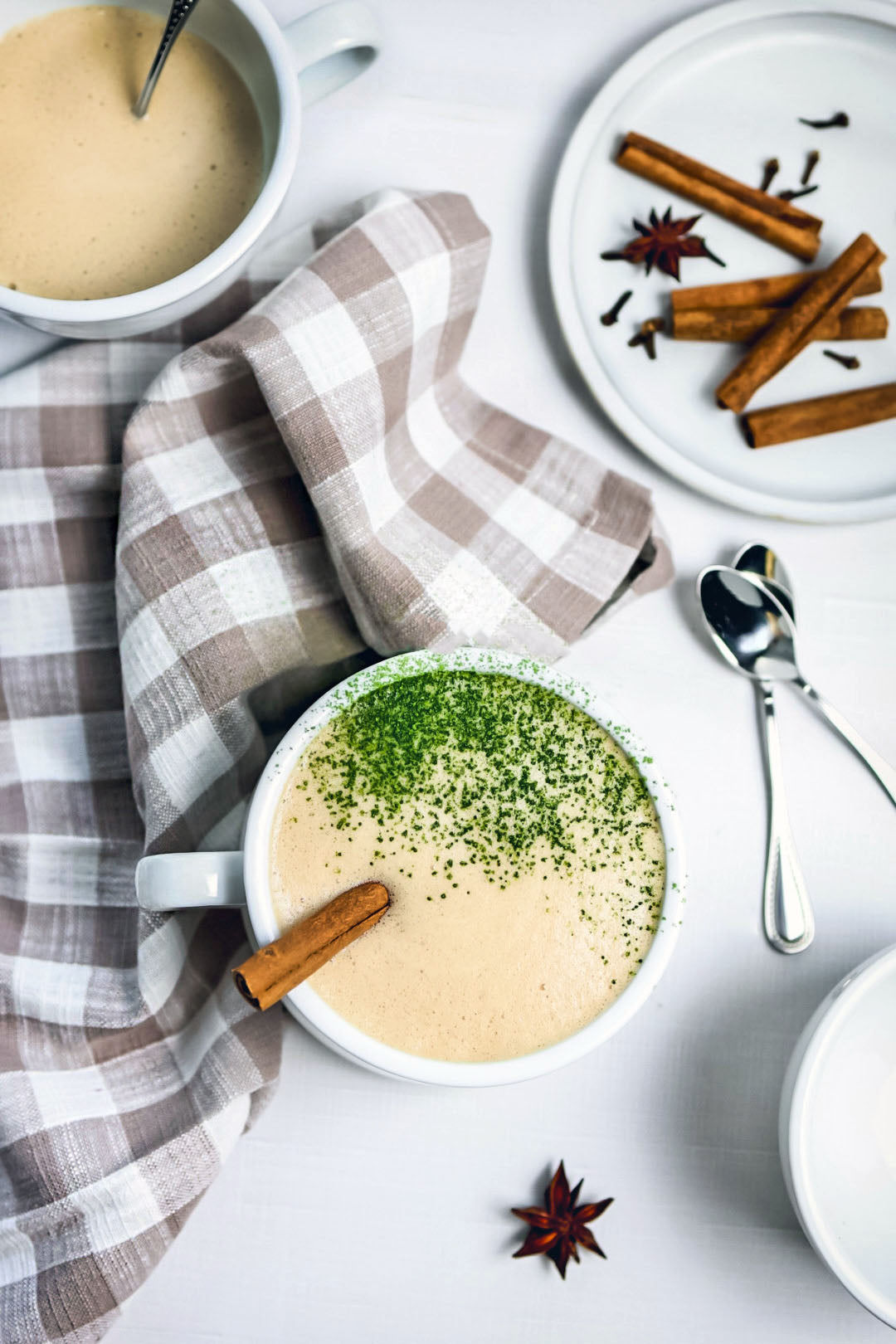 A cozy Matcha Chai Latte in a white mug, topped with vibrant green matcha and a cinnamon stick, surrounded by chai spices and a soft plaid napkin.