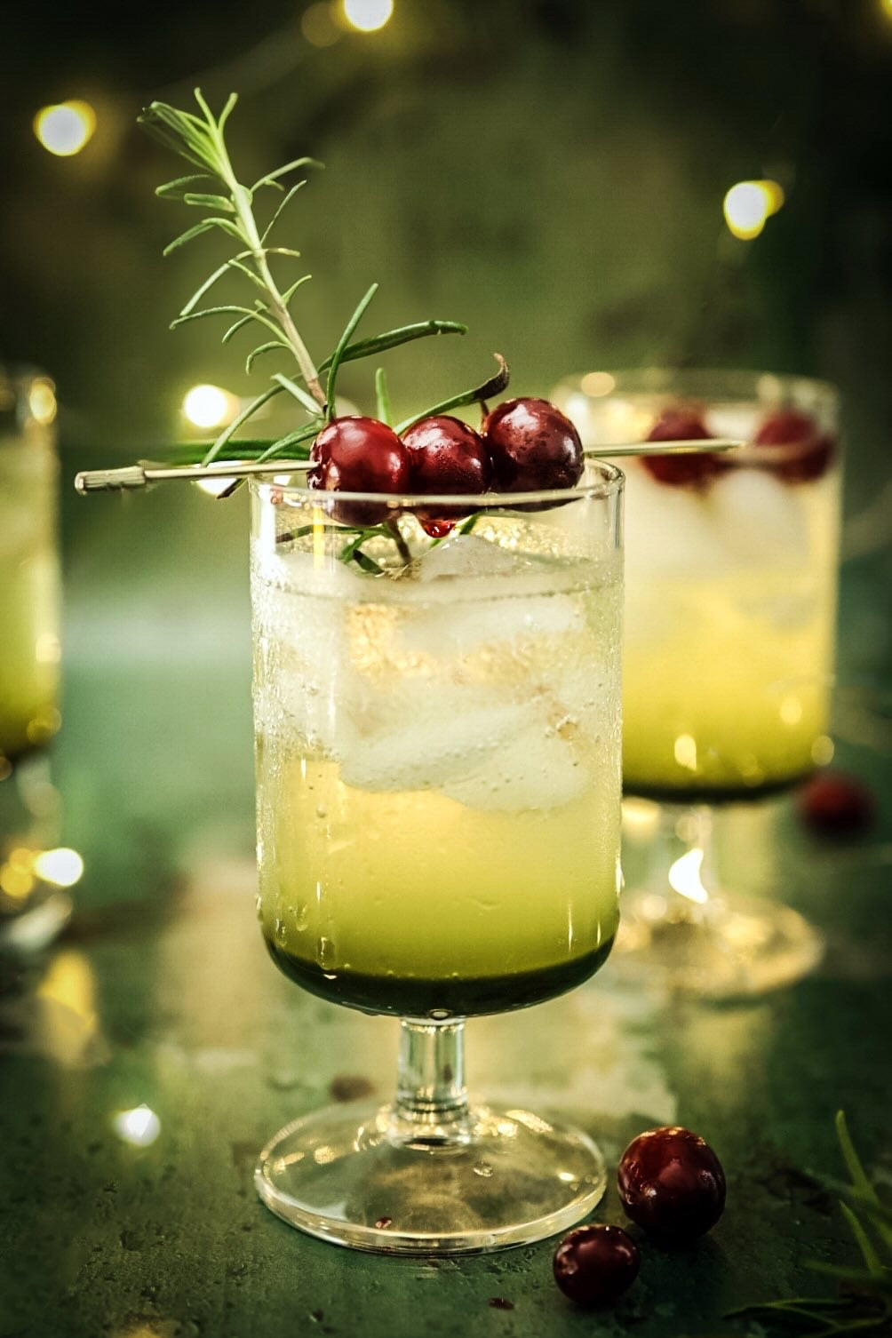 A festive Cranberry Matcha Mocktail in a glass garnished with fresh cranberries and a sprig of rosemary, set against twinkling holiday lights.