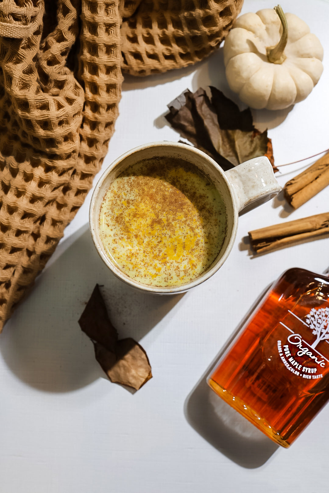 A cozy Fall Spice Matcha Latte in a rustic ceramic mug, sprinkled with cinnamon, surrounded by a soft brown knit blanket, cinnamon sticks, and a white pumpkin.