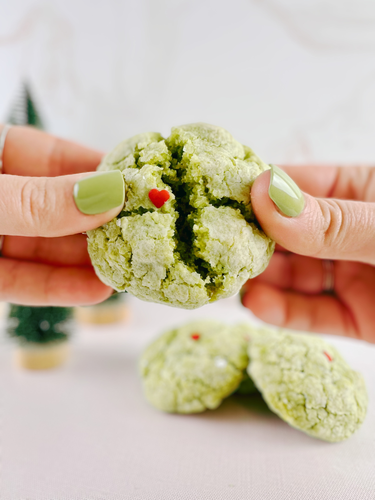 Close-up of hands breaking apart a vibrant green Matcha Grinch Cookie with a tiny red heart sprinkle, set against a festive holiday background.