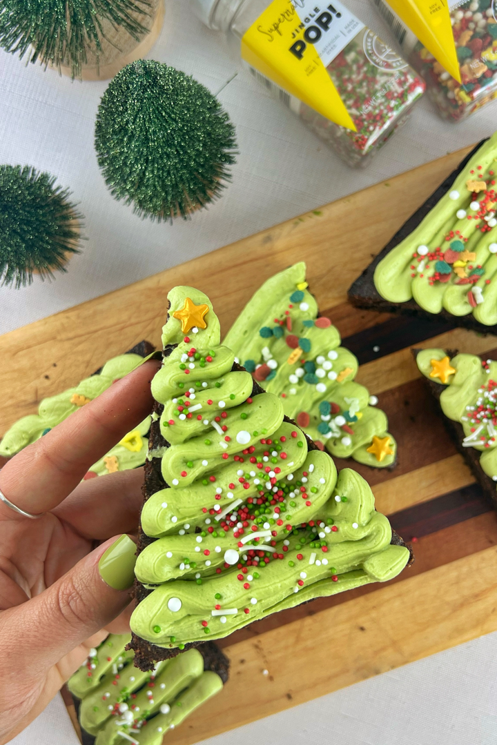 Hand holding a triangle-shaped Matcha Brownie Christmas Tree topped with green matcha frosting, holiday sprinkles, and a golden star, with festive decorations in the background.