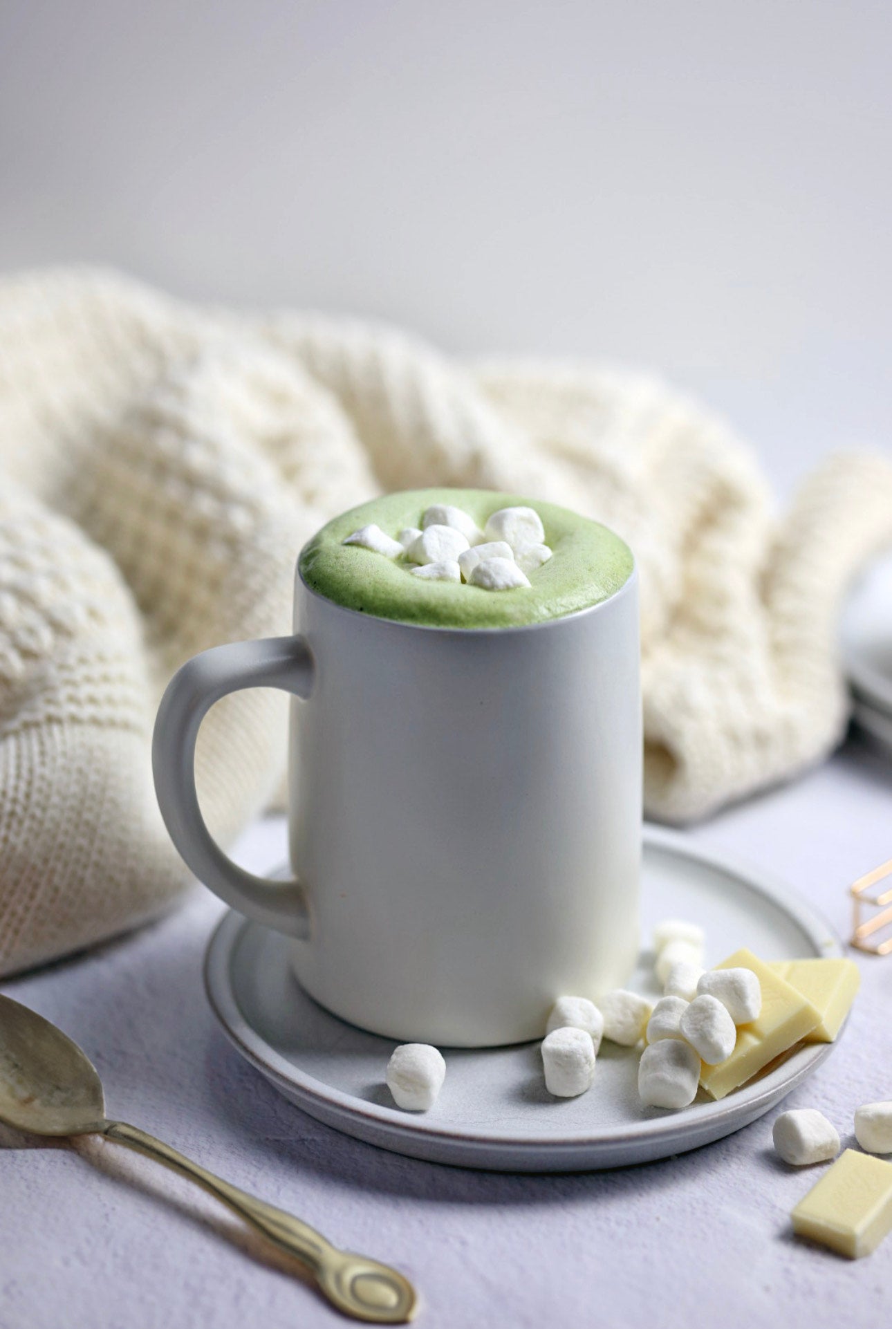 A cozy mug of Matcha White Hot Chocolate topped with mini marshmallows, surrounded by a soft cream blanket, white chocolate chunks, and a golden spoon