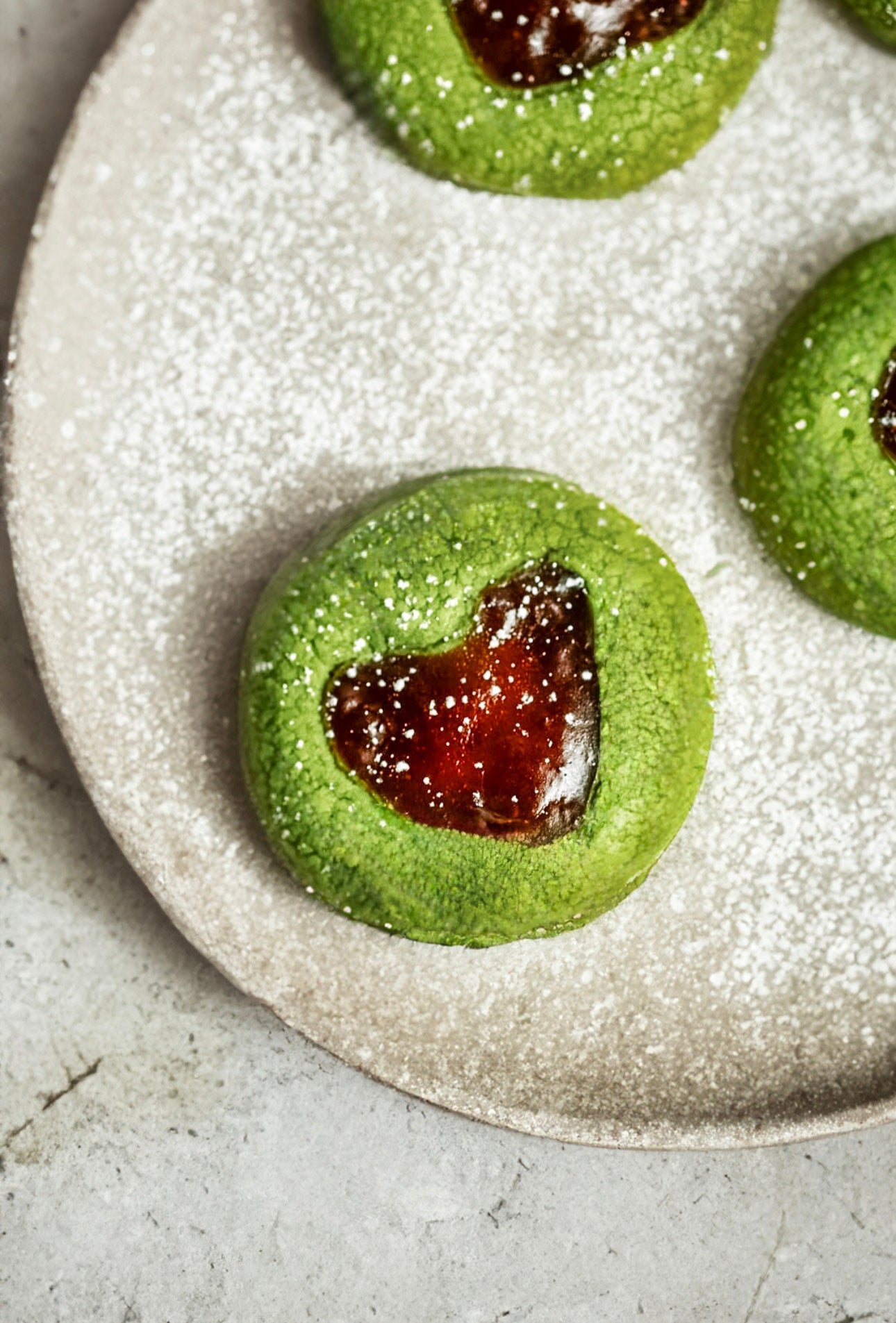 Heart-shaped Matcha Thumbprint Cookie filled with strawberry jam, dusted with powdered sugar on a neutral plate.