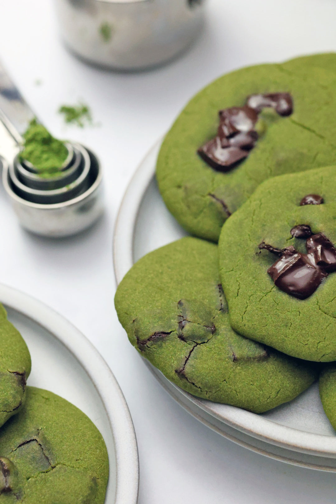 A plate of chewy green Mochi Matcha Cookies topped with gooey chocolate chunks, with a measuring spoon filled with vibrant matcha powder in the background
