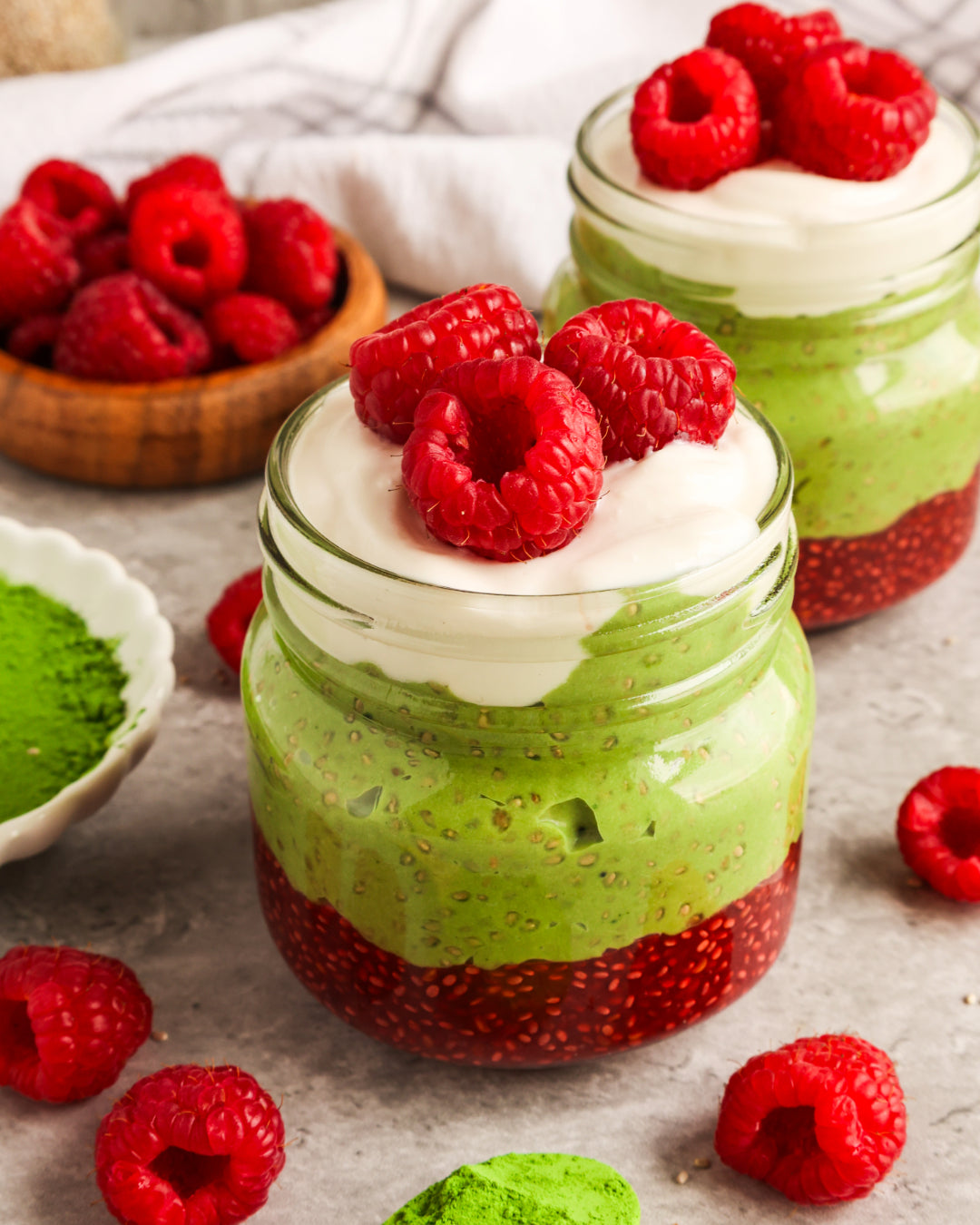 Layered Raspberry Matcha Chia Pudding in glass jars topped with creamy yogurt and fresh raspberries, surrounded by scattered matcha powder and raspberries.