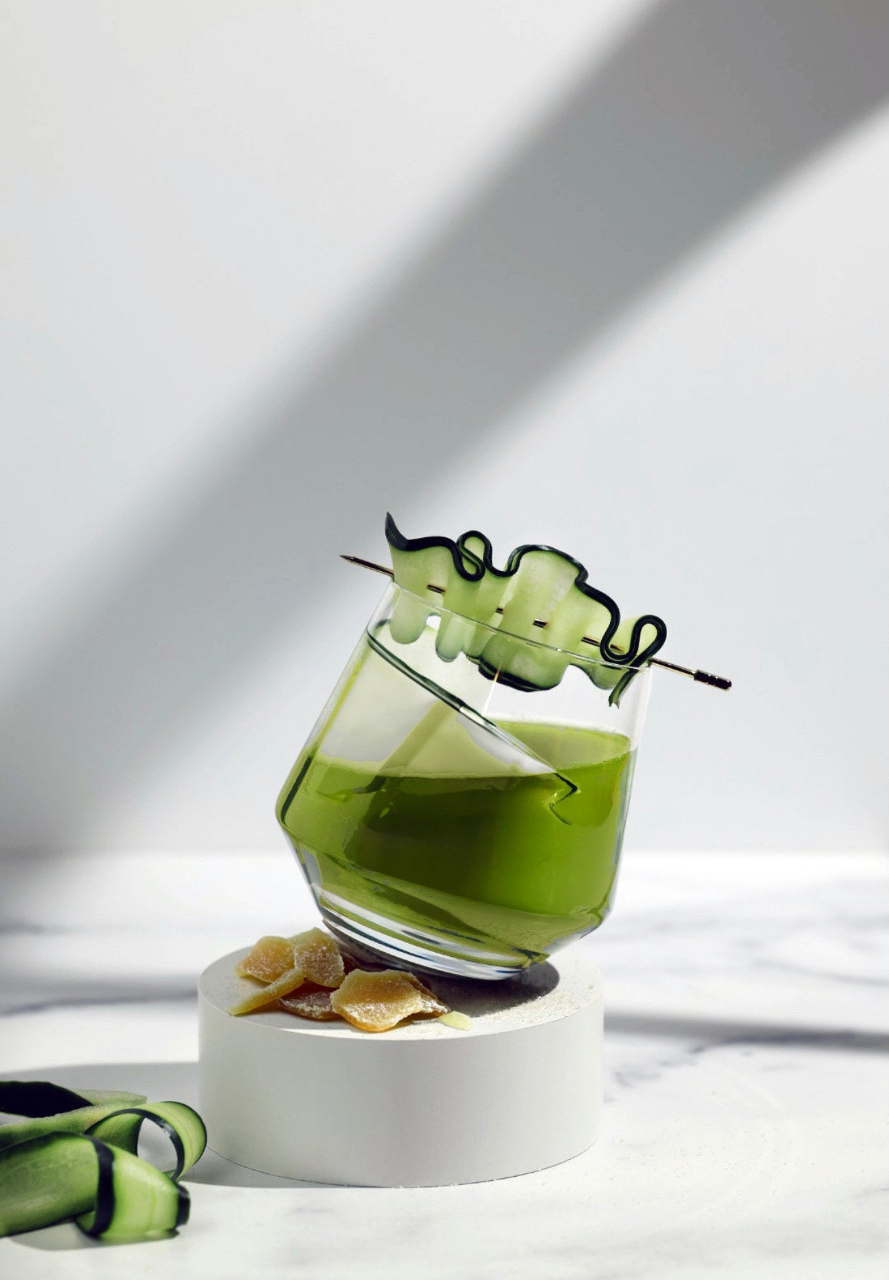 Emerald Elixir Matcha Cocktail in a tilted glass, garnished with a cucumber ribbon and surrounded by candied ginger slices on a white pedestal