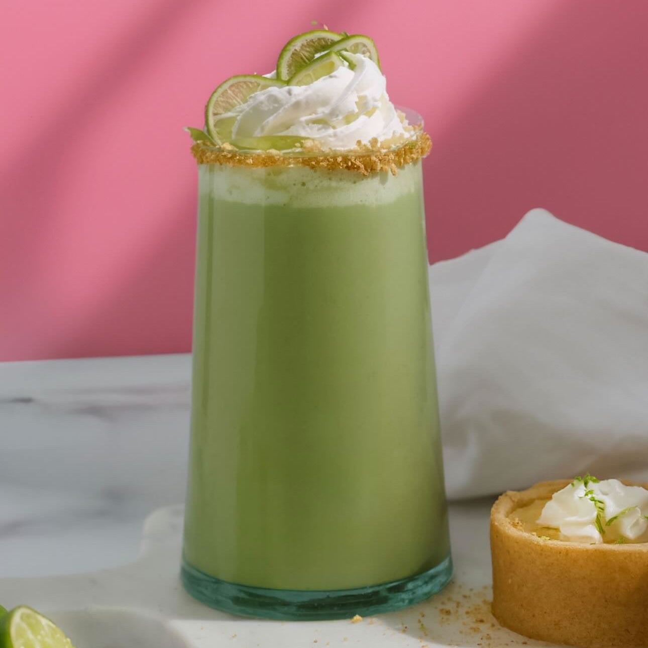 A tall glass of Key Lime Pie Matcha topped with whipped cream, lime slices, and a golden rim garnish, sitting on a marble board with a small key lime pie beside it.