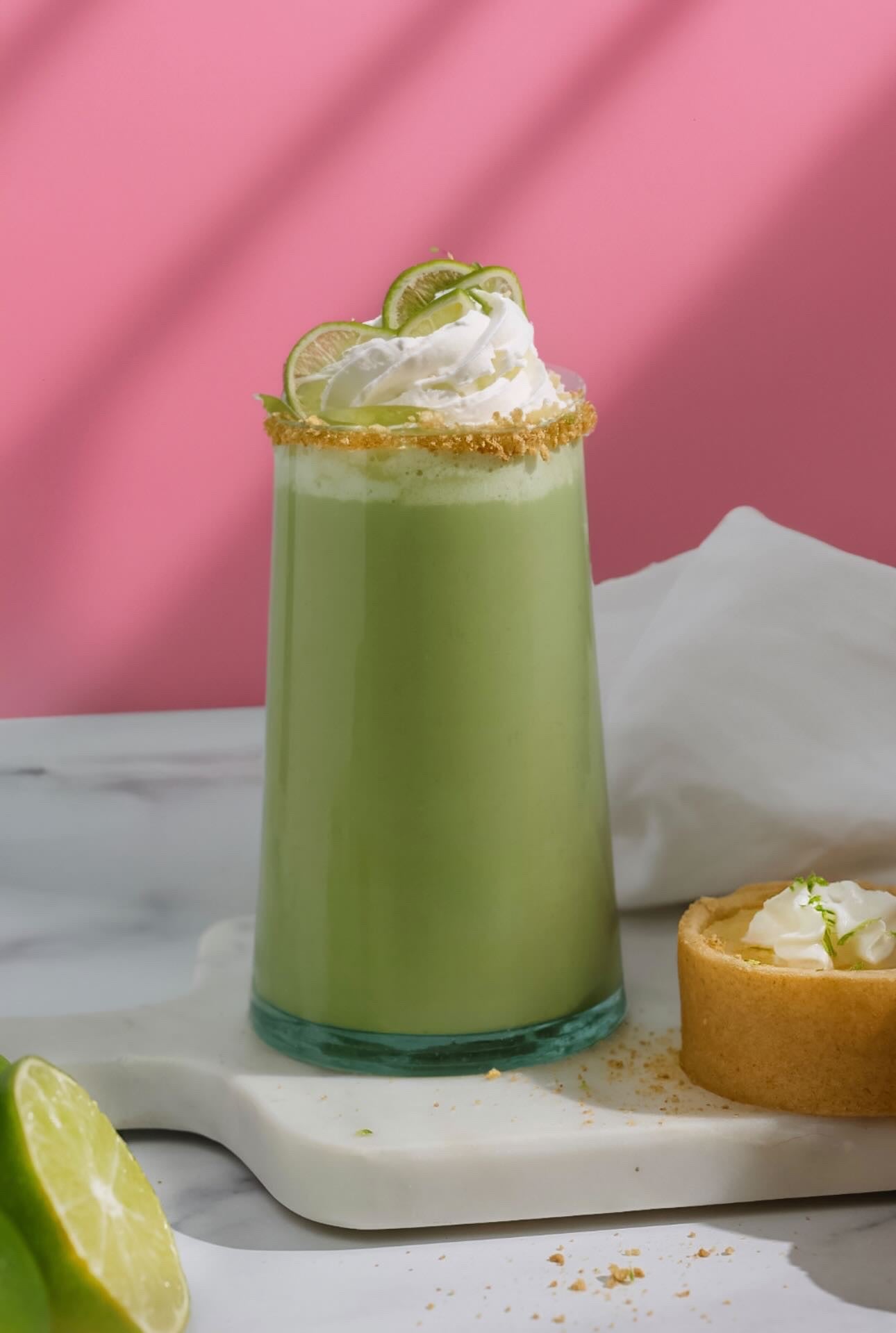A tall glass of Key Lime Pie Matcha topped with whipped cream, lime slices, and a golden rim garnish, sitting on a marble board with a small key lime pie beside it.