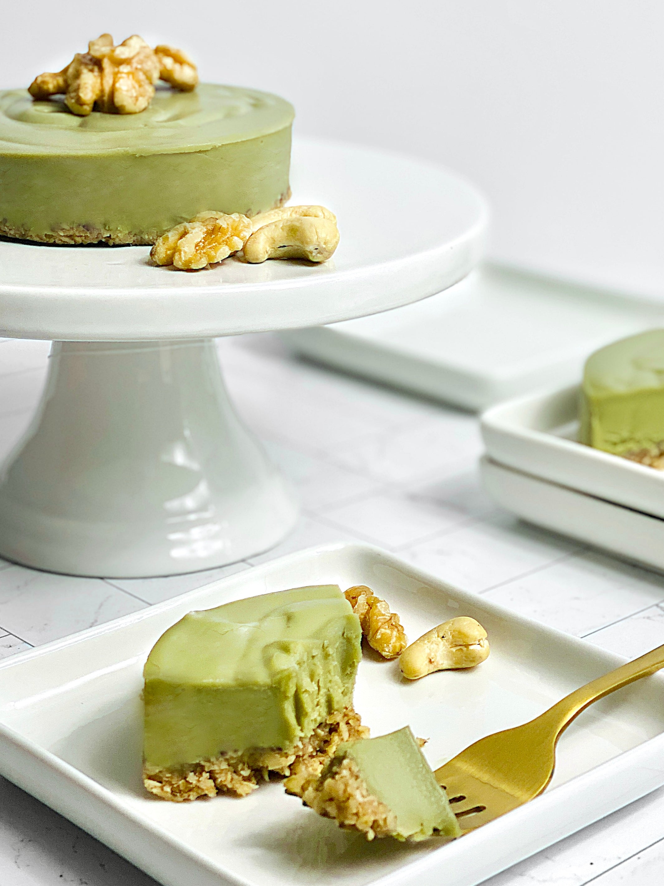A creamy Vegan Matcha Cheesecake on a white cake stand, topped with a walnut. A slice sits on a white plate with a golden fork, accompanied by cashews and walnuts.