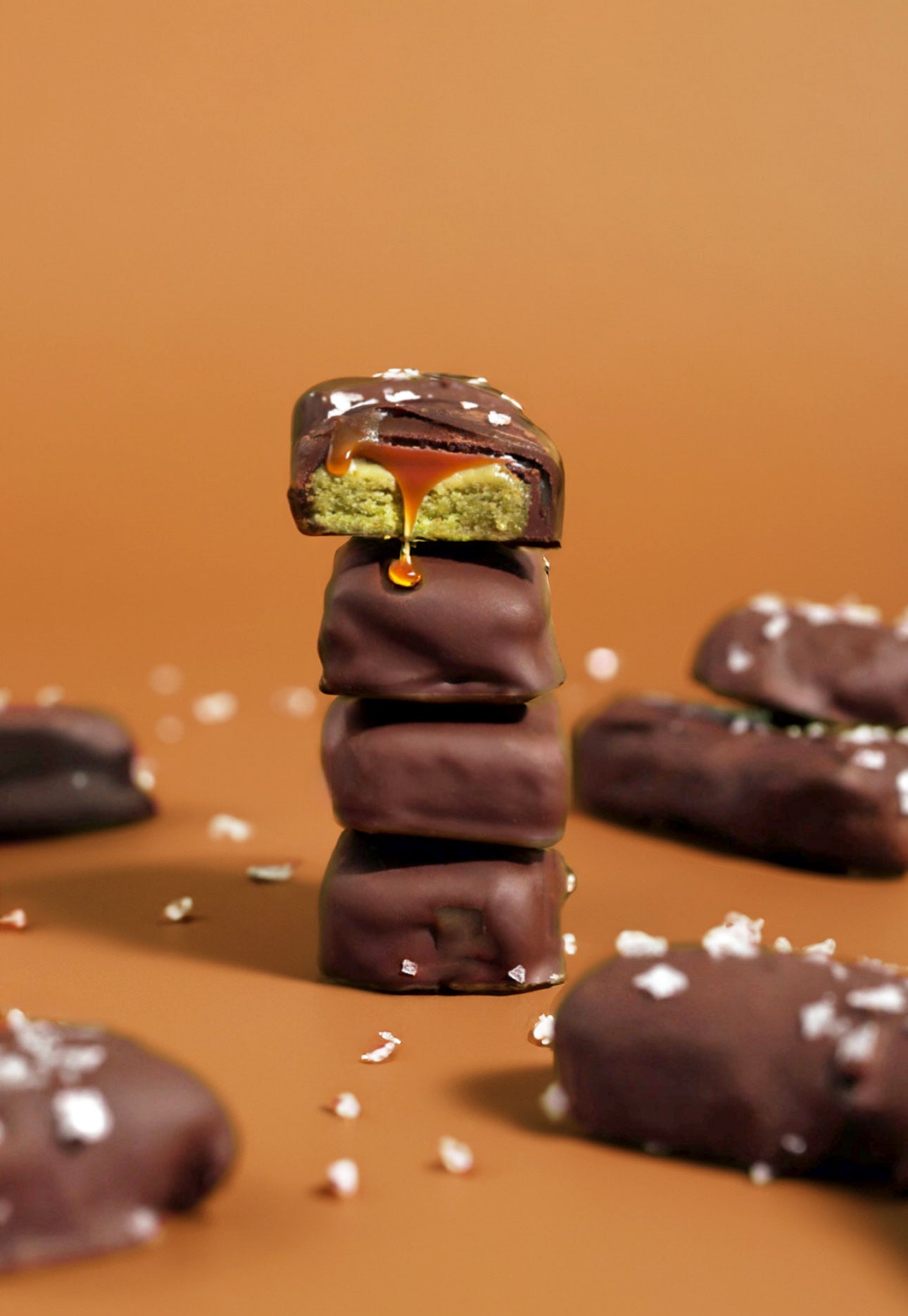 A stack of four Healthy-ish Matcha Twix Bars covered in smooth chocolate, with the top bar cut open revealing a gooey caramel drip and vibrant green matcha shortbread, sprinkled with sea salt flakes.