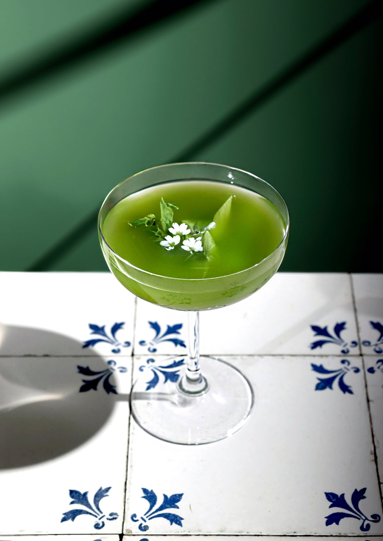 Elegant Matcha Martini in a coupe glass, garnished with fresh mint and edible flowers, set on a white and blue tiled surface with a moody green background.