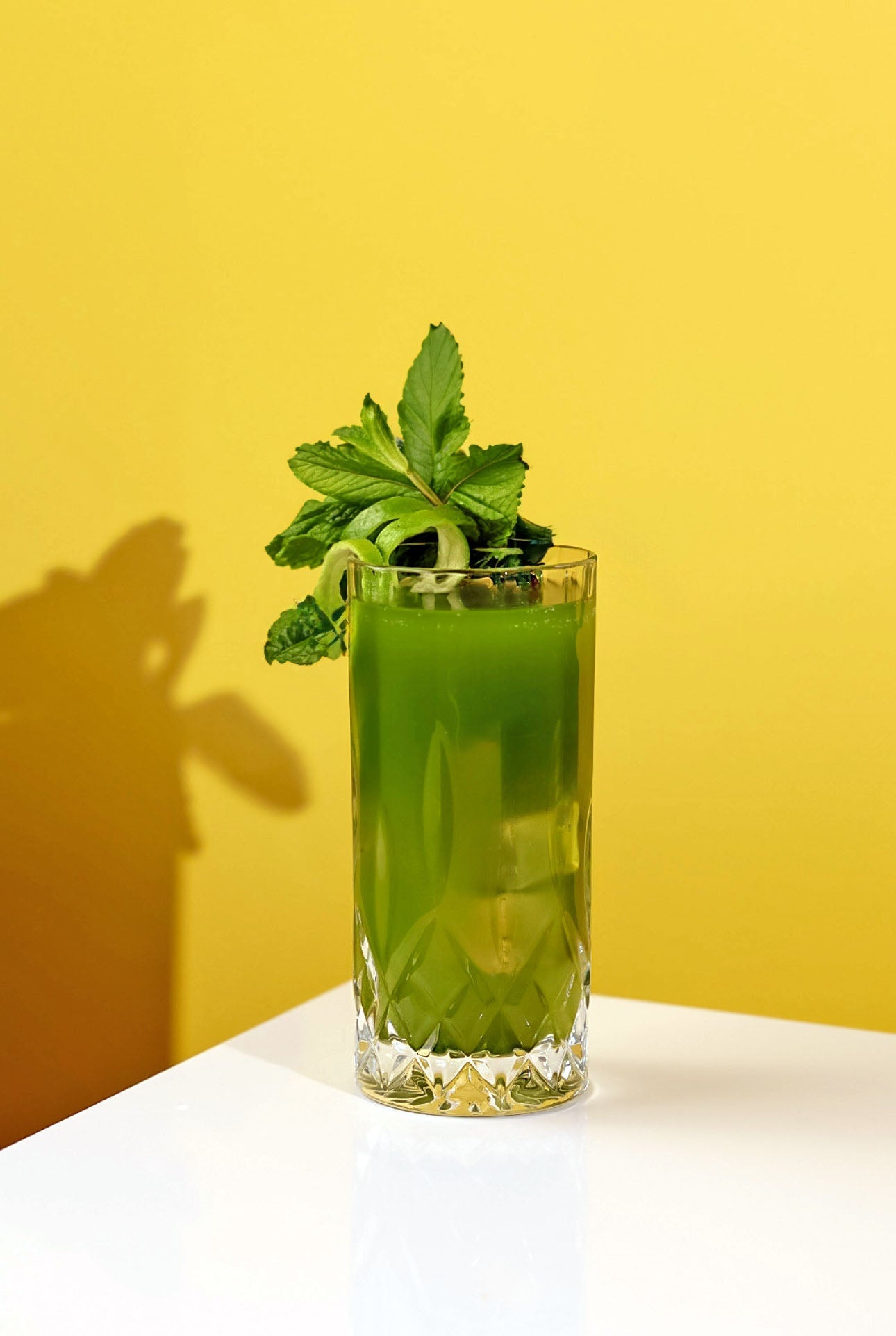 Vibrant green Matcha Mojito Mocktail in a tall glass, garnished with fresh mint and a lime peel, set against a bright yellow background.