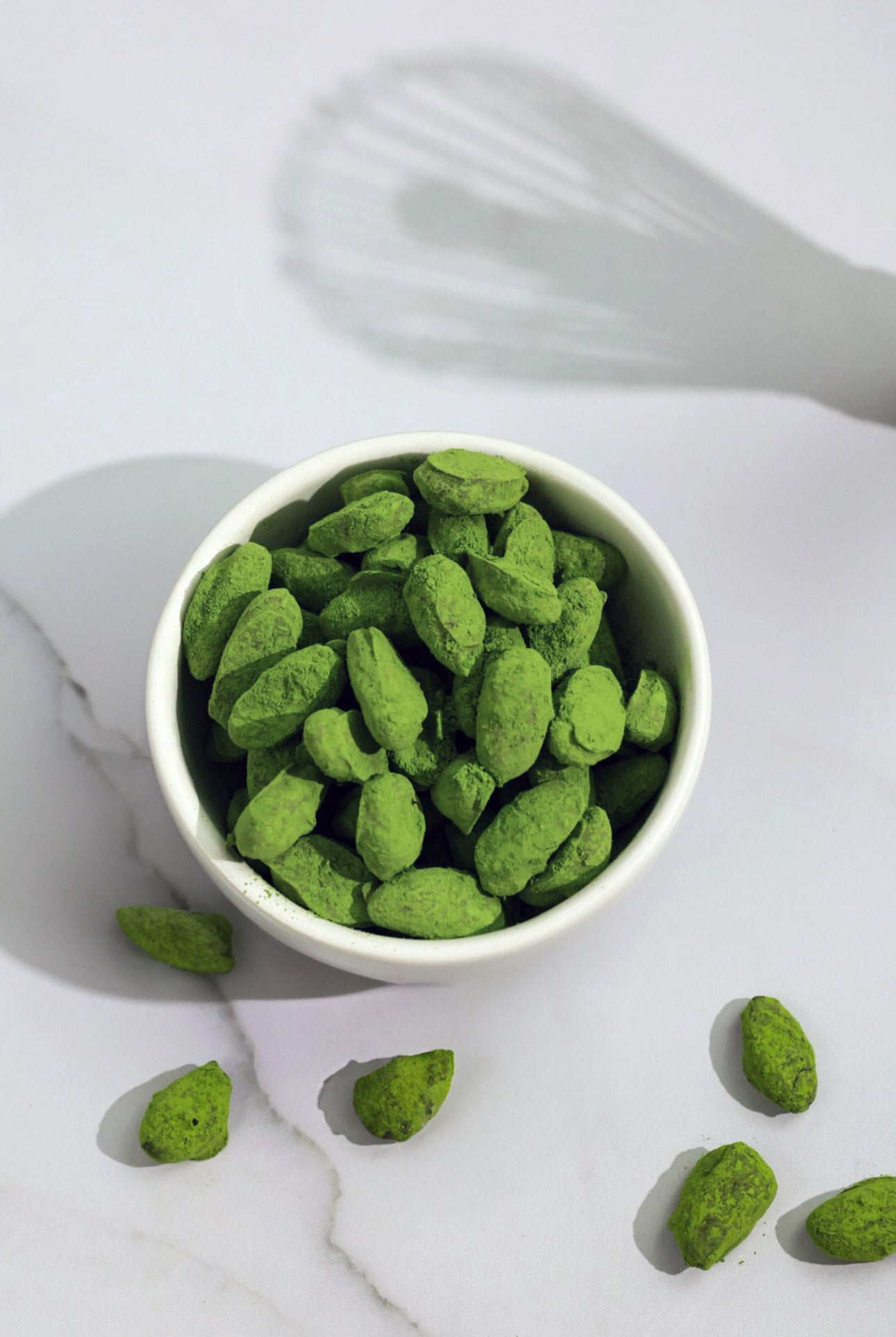 A white bowl filled with chocolate-covered almonds dusted in vibrant green matcha powder, set against a clean white background with scattered almonds around.