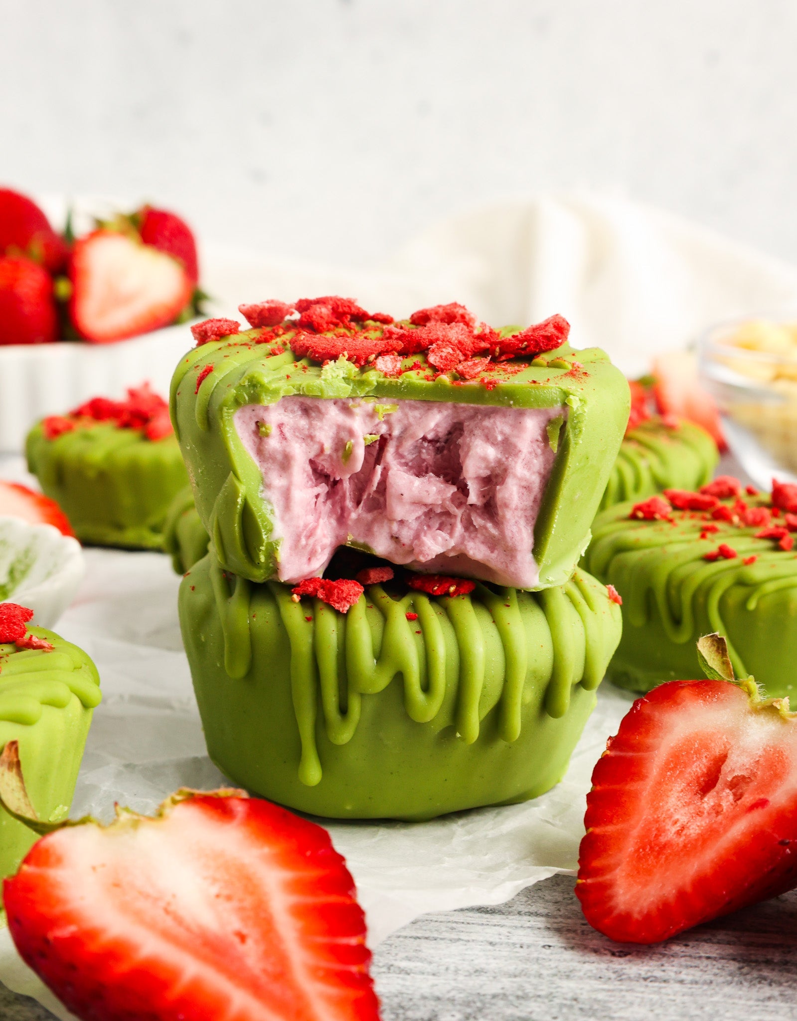 Frozen strawberry yogurt cups coated in vibrant matcha white chocolate, garnished with freeze-dried strawberries, and surrounded by fresh strawberry slices.
