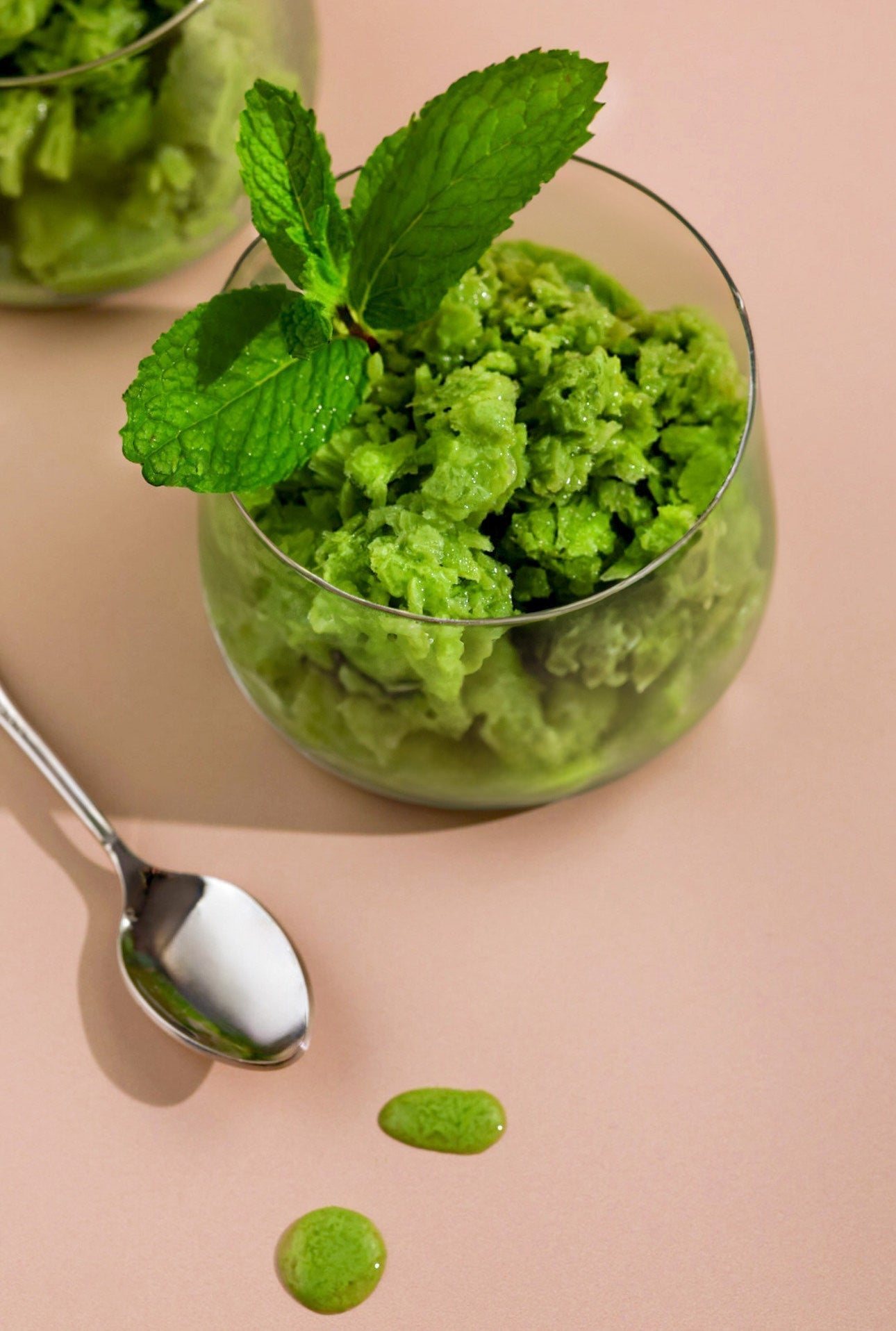 A glass filled with vibrant green Matcha Granita, topped with fresh mint leaves, placed on a soft pink background with a spoon nearby and a few drops of melted granita.