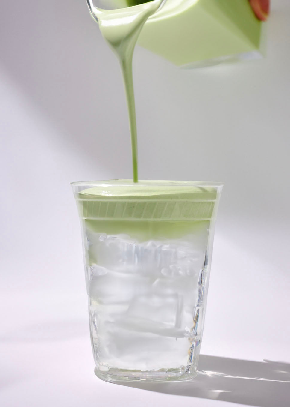 A glass filled with iced coconut water, topped with creamy matcha cold foam being poured in, set against a bright, clean background.