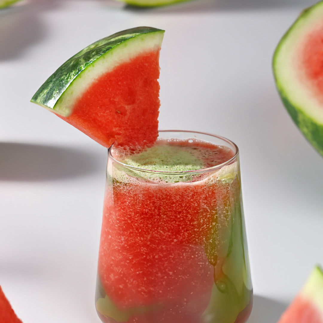 A vibrant glass of Watermelon Matcha Agua Fresca Smoothie garnished with a fresh watermelon slice, surrounded by watermelon wedges on a clean white background.