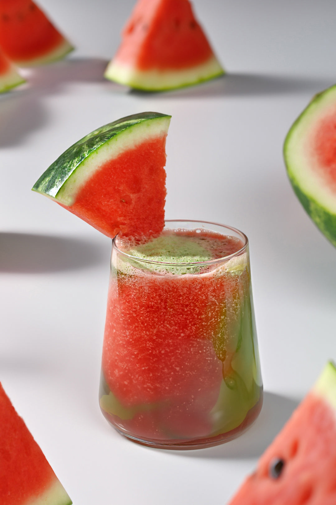 A vibrant glass of Watermelon Matcha Agua Fresca Smoothie garnished with a fresh watermelon slice, surrounded by watermelon wedges on a clean white background.